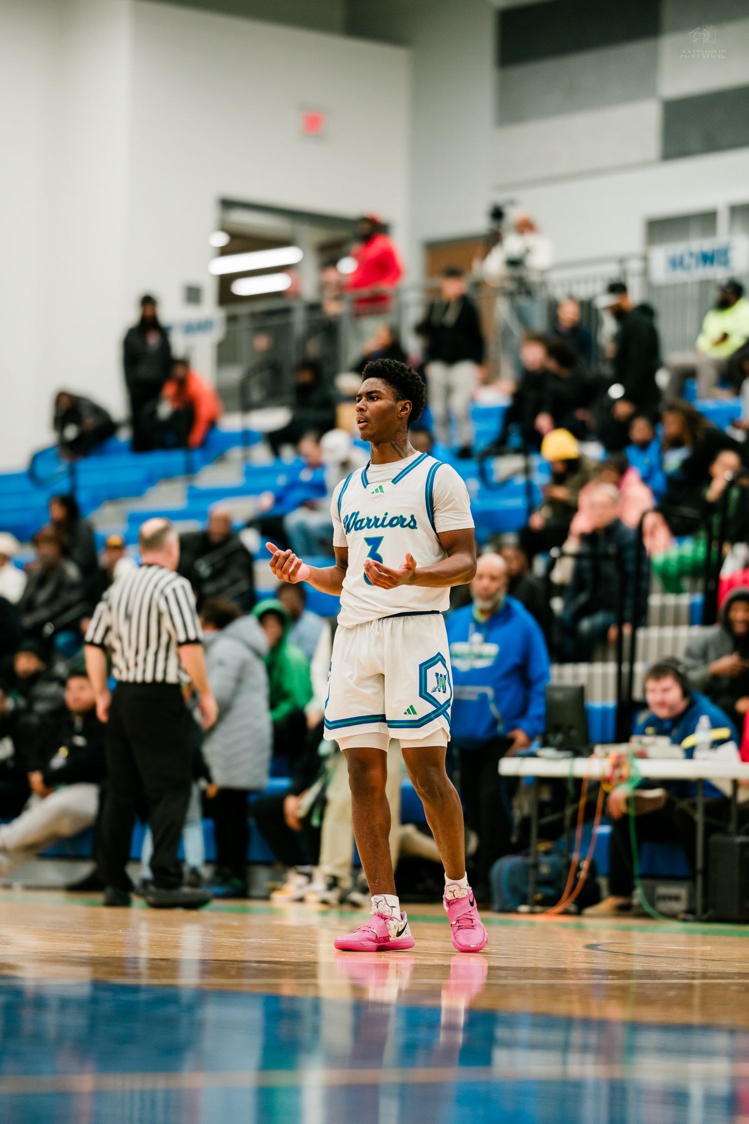 Warriors player number 3 walks the court in pink sneakers during a high school basketball game with fans watching.