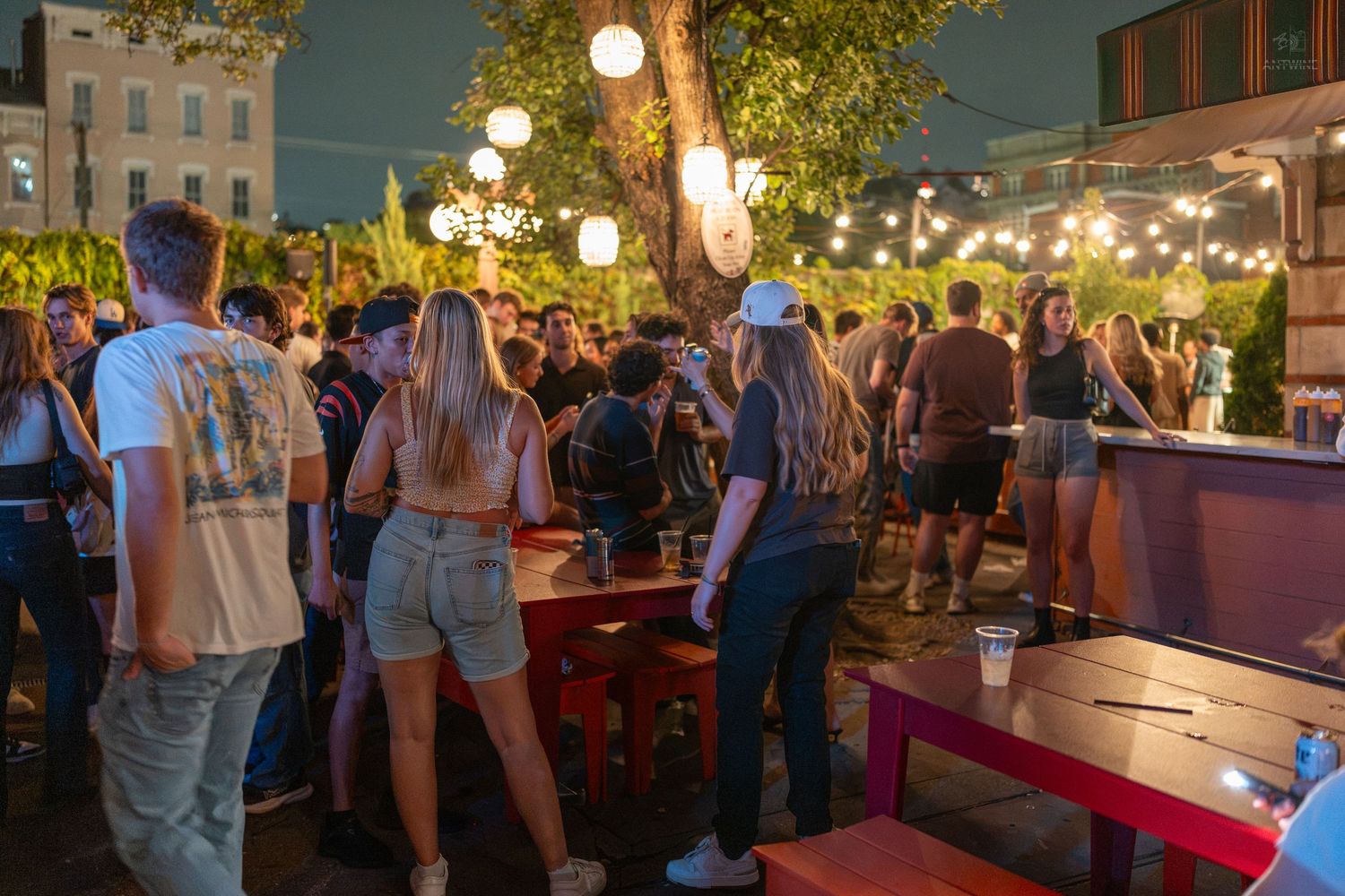 Crowded outdoor evening bar event with string lights, trees, and colorful furniture in a lively urban setting.
