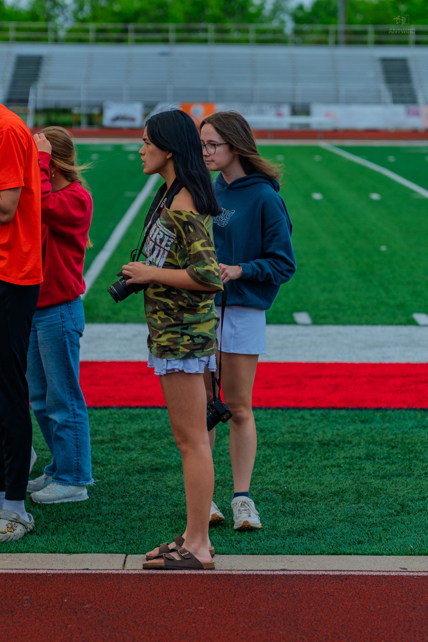 Two young women holding cameras stand on a red track beside a green football field during an outdoor event.