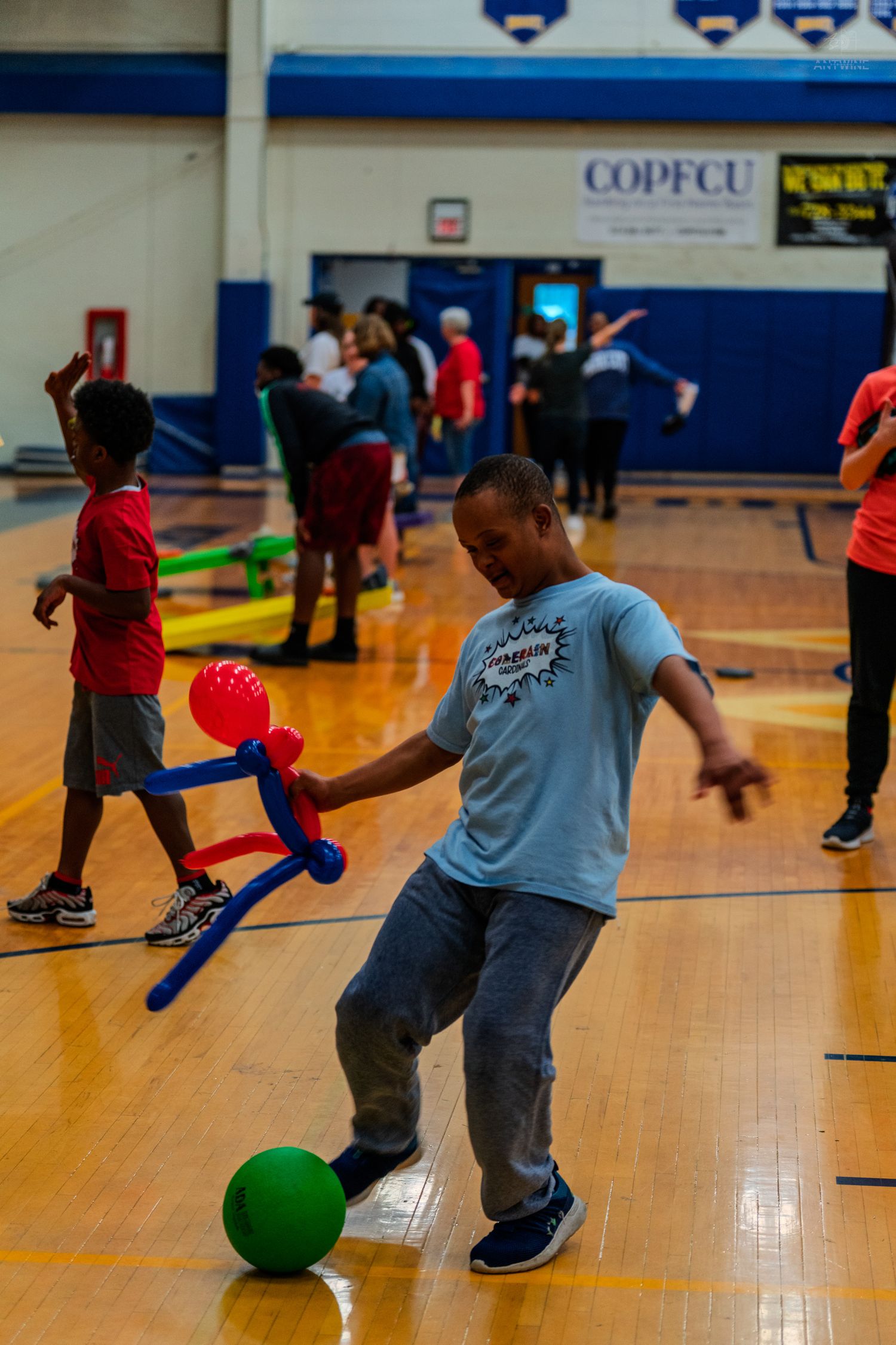 Children playing indoor sports in a gymnasium, with a boy kicking a green ball during a fun recreational activity.