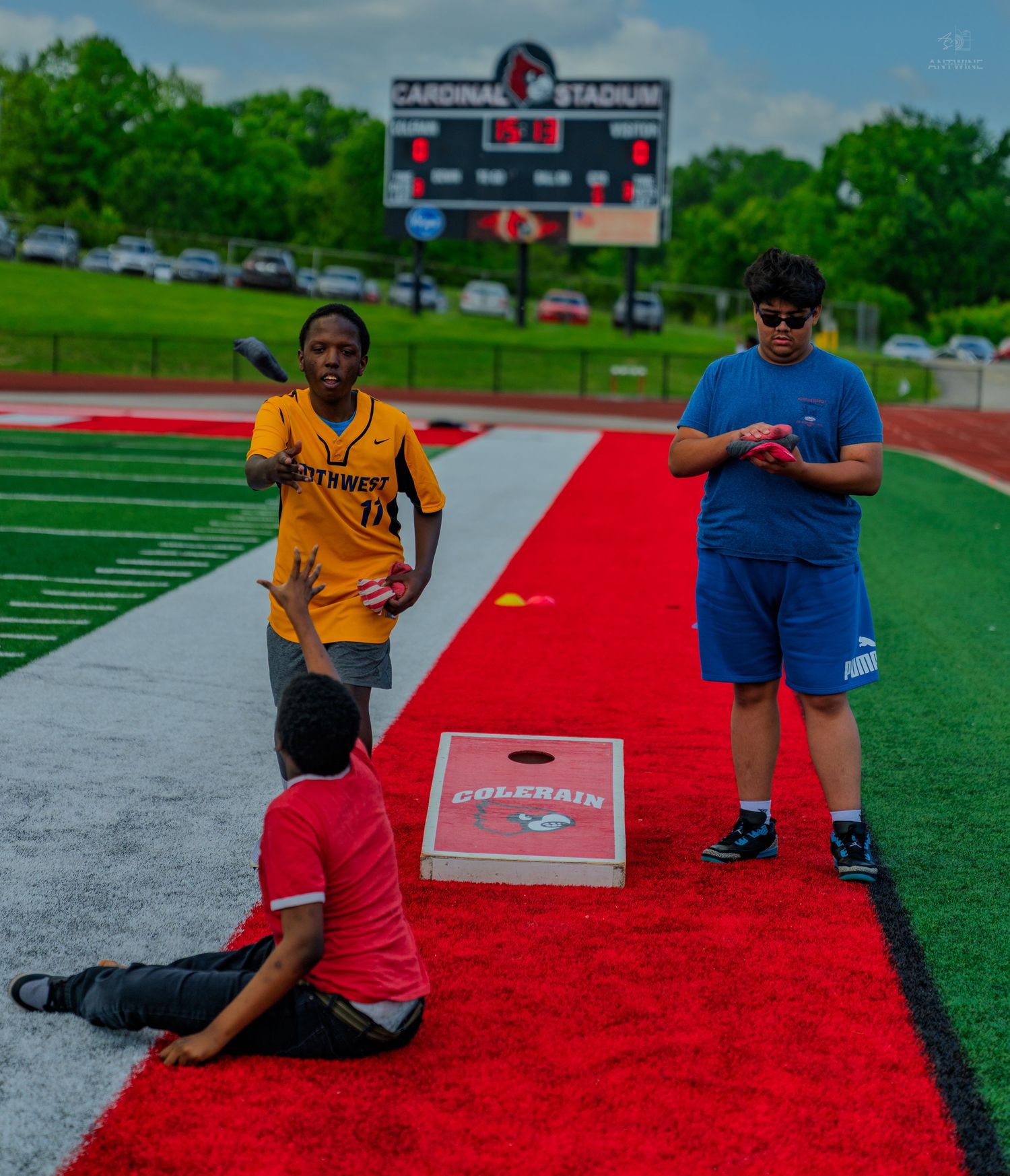 Three kids playing cornhole on a red turf track at Cardinal Stadium on a cloudy day.