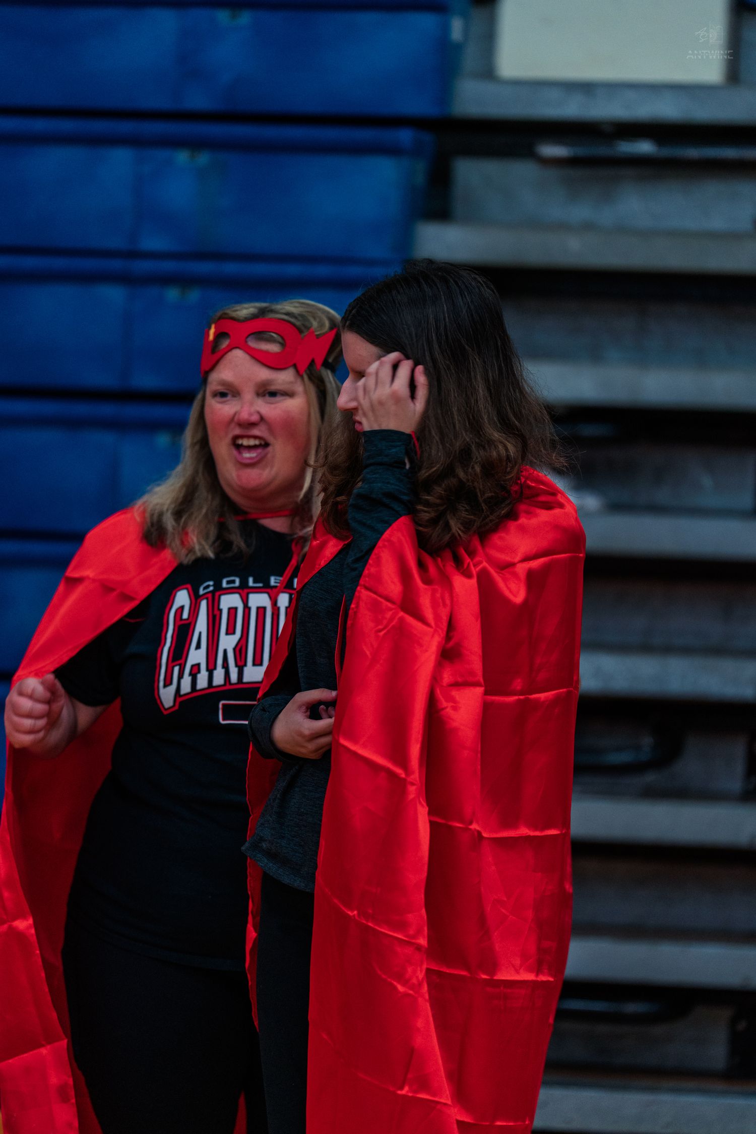 Two women in red capes and masks cheering at a Cardinals school sports event in bleachers.