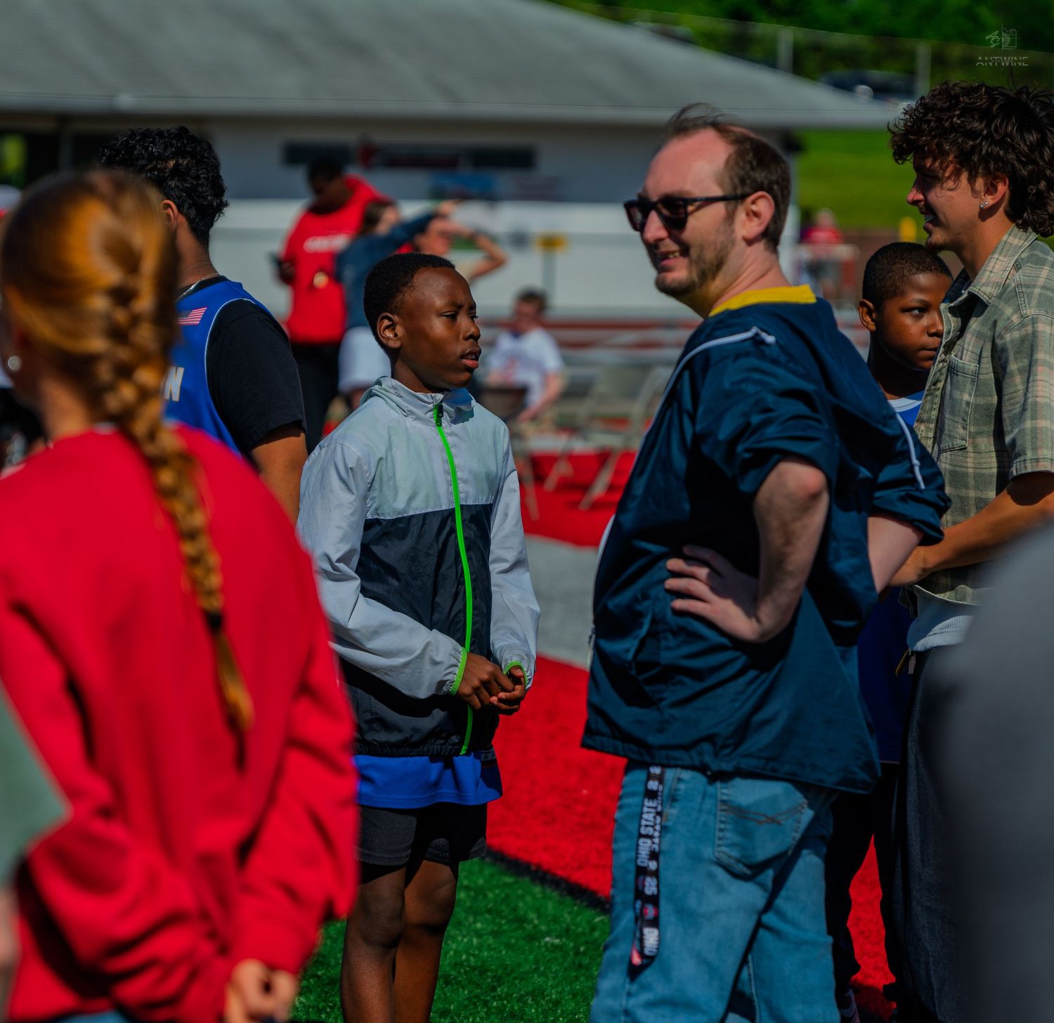 Adults and children gather outdoors at a youth track and field event on a green sports field.