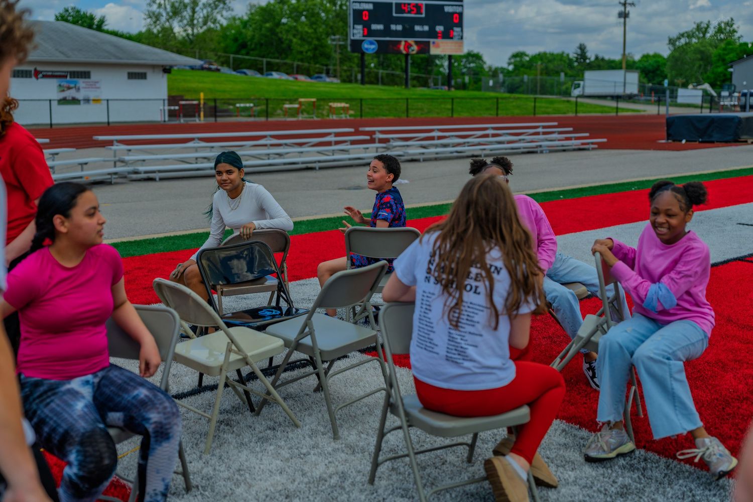 Girls relaxing in folding chairs on a red track near stadium bleachers during an outdoor sports event.