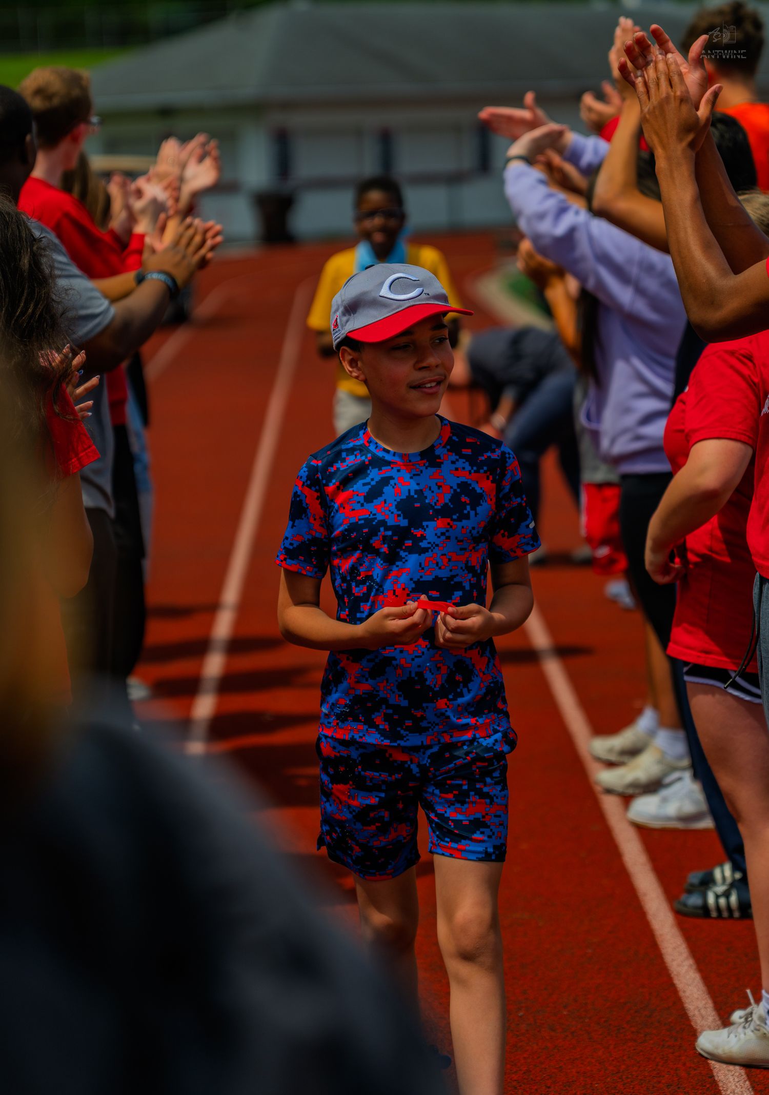Young boy in blue patterned shirt and red cap walks through a high-five tunnel on a red running track.