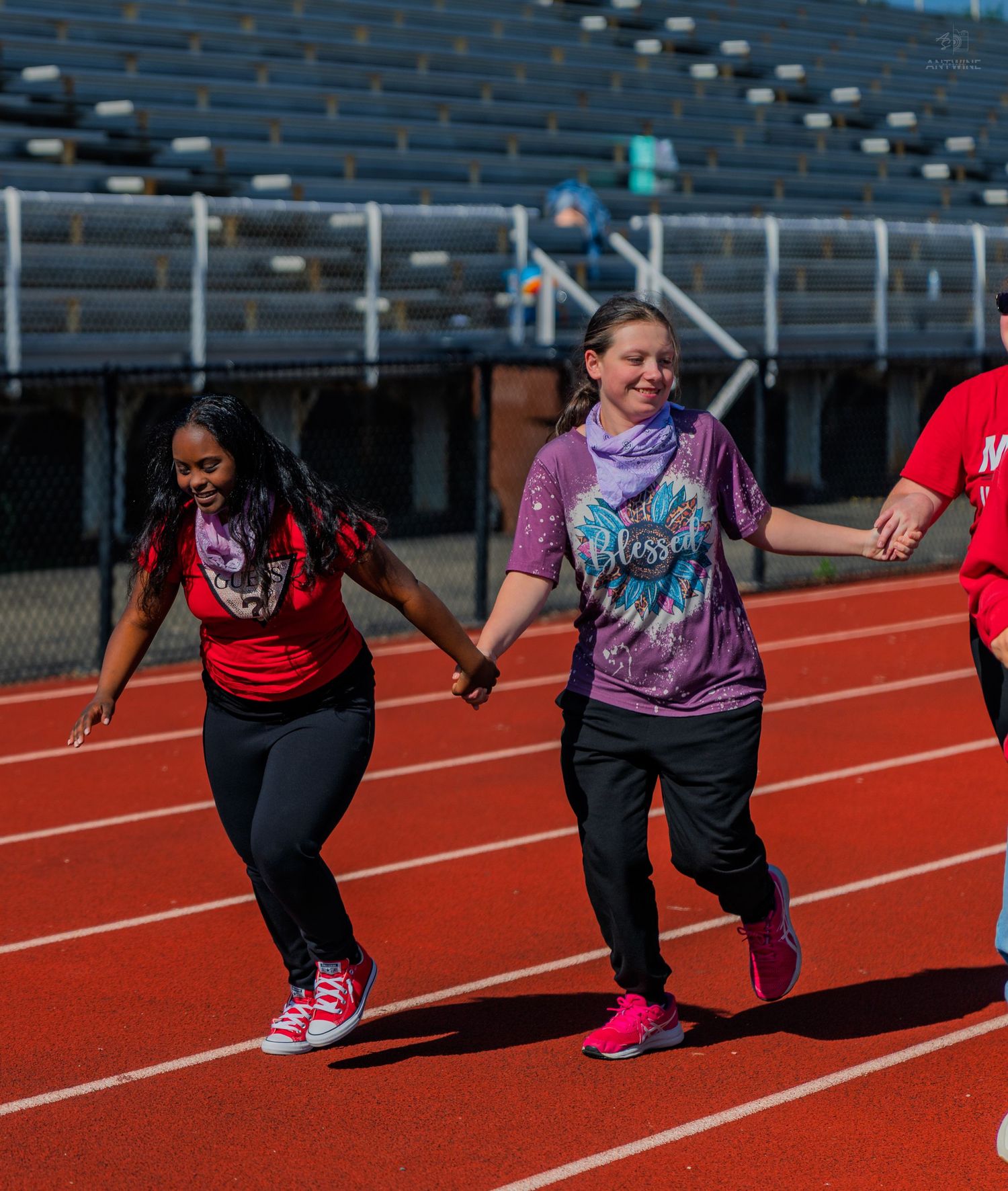 Three girls hold hands while running together on a red track during an outdoor school sports event.