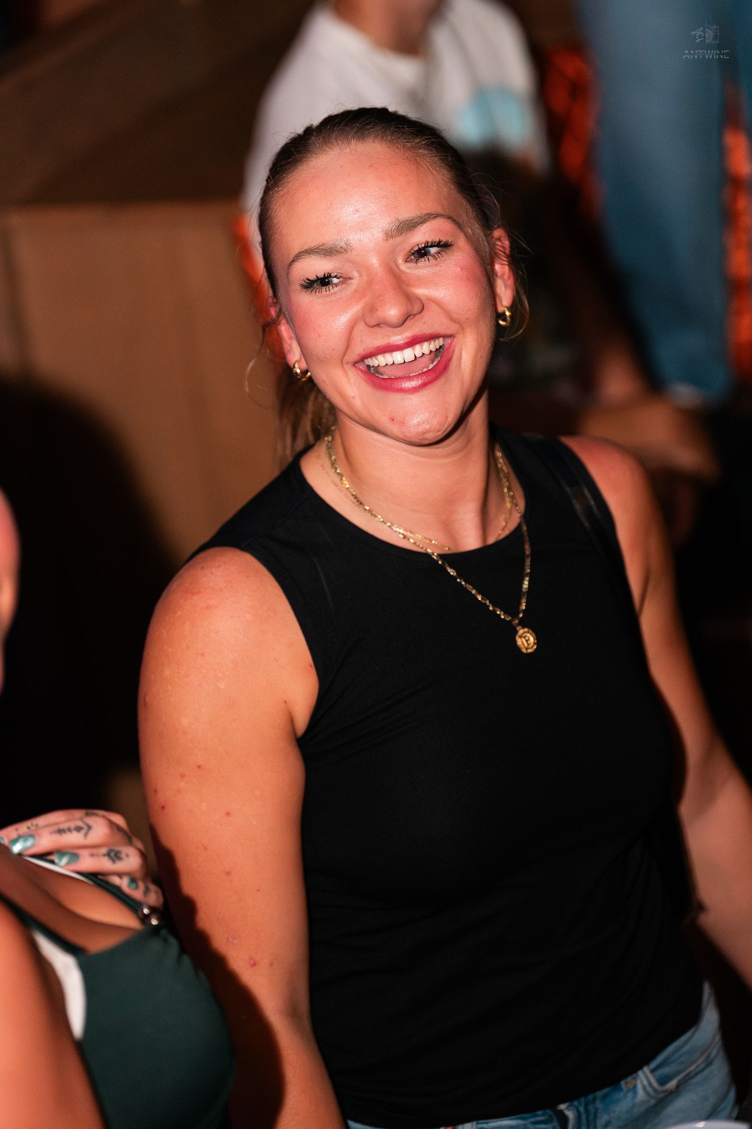 Smiling young woman in black sleeveless top and gold necklace at a social gathering.