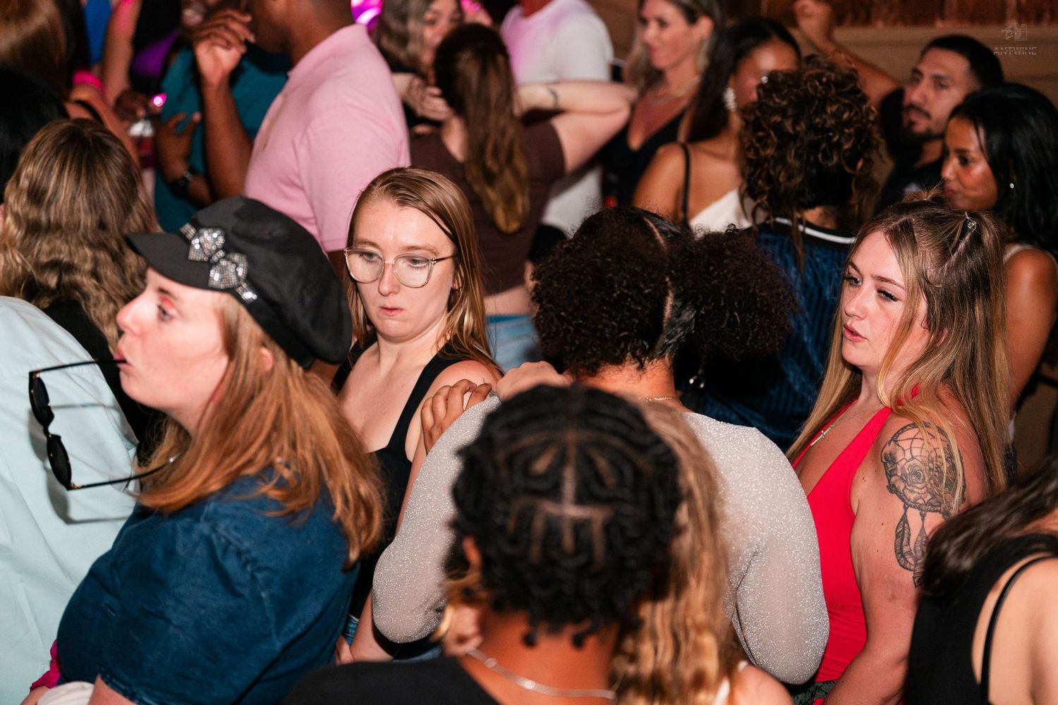 Diverse group of young adults at a packed nightclub event, woman in red dress with tattoo visible.