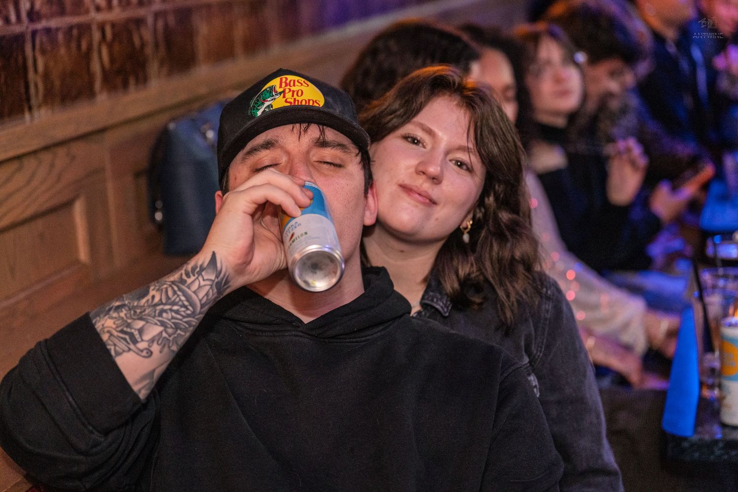 Tattooed man drinking a canned beverage while a woman leans on his shoulder at a bar.