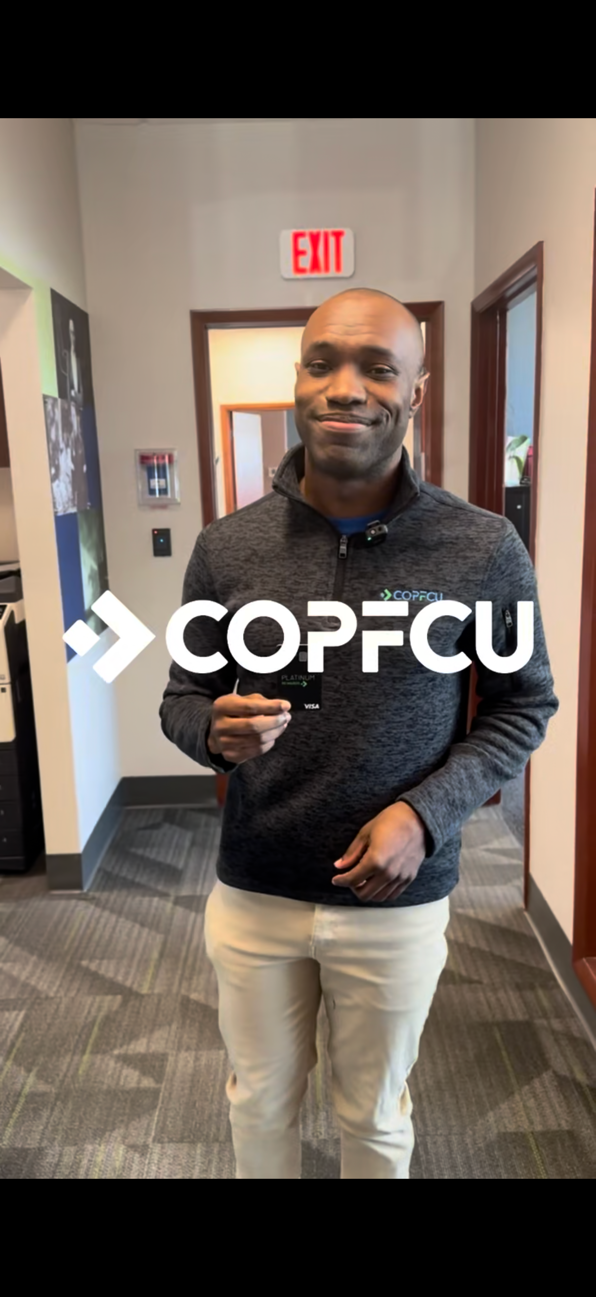 A smiling man in a COPFCU branded gray pullover holds a card inside a credit union office hallway.
