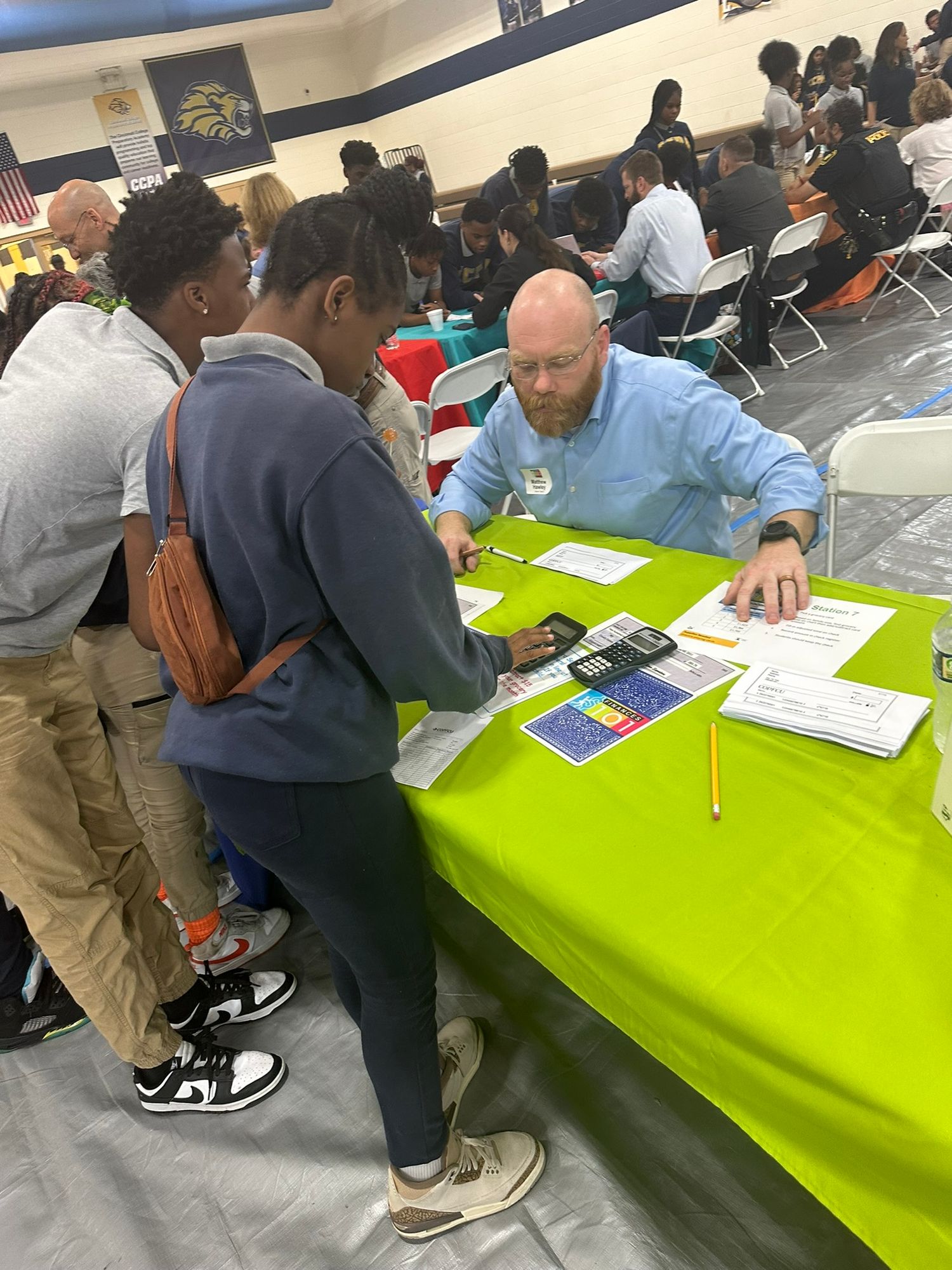 A bearded teacher helps two students with a calculator at a green table during an educational school event.