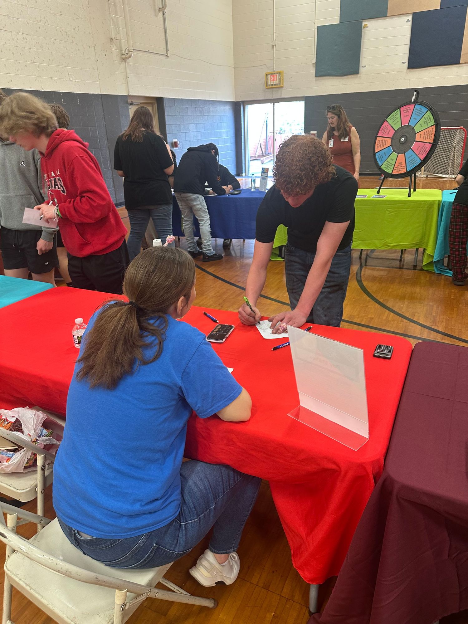 People gathered at indoor school event with red tables, a colorful prize wheel, and participants signing forms in a gym.