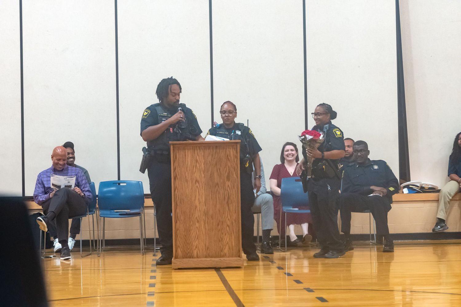 Law enforcement officers speak at a community meeting podium inside a school gymnasium with attendees seated nearby.