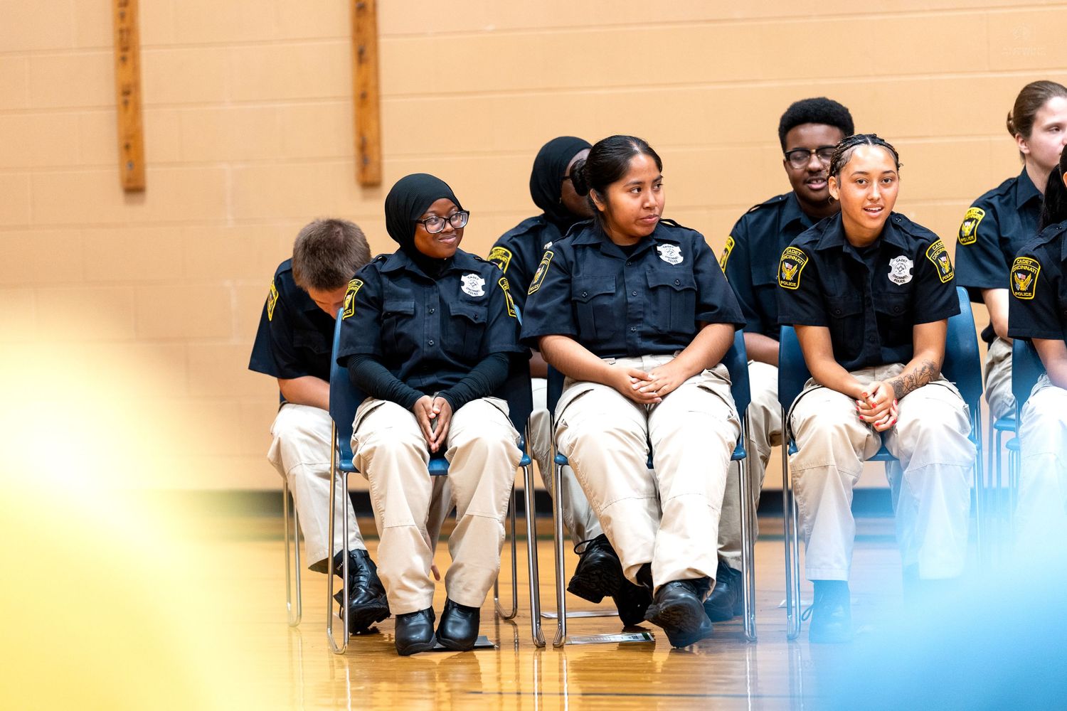 Young students in matching navy uniforms and khaki pants sit in chairs during a school ceremony in a gymnasium.