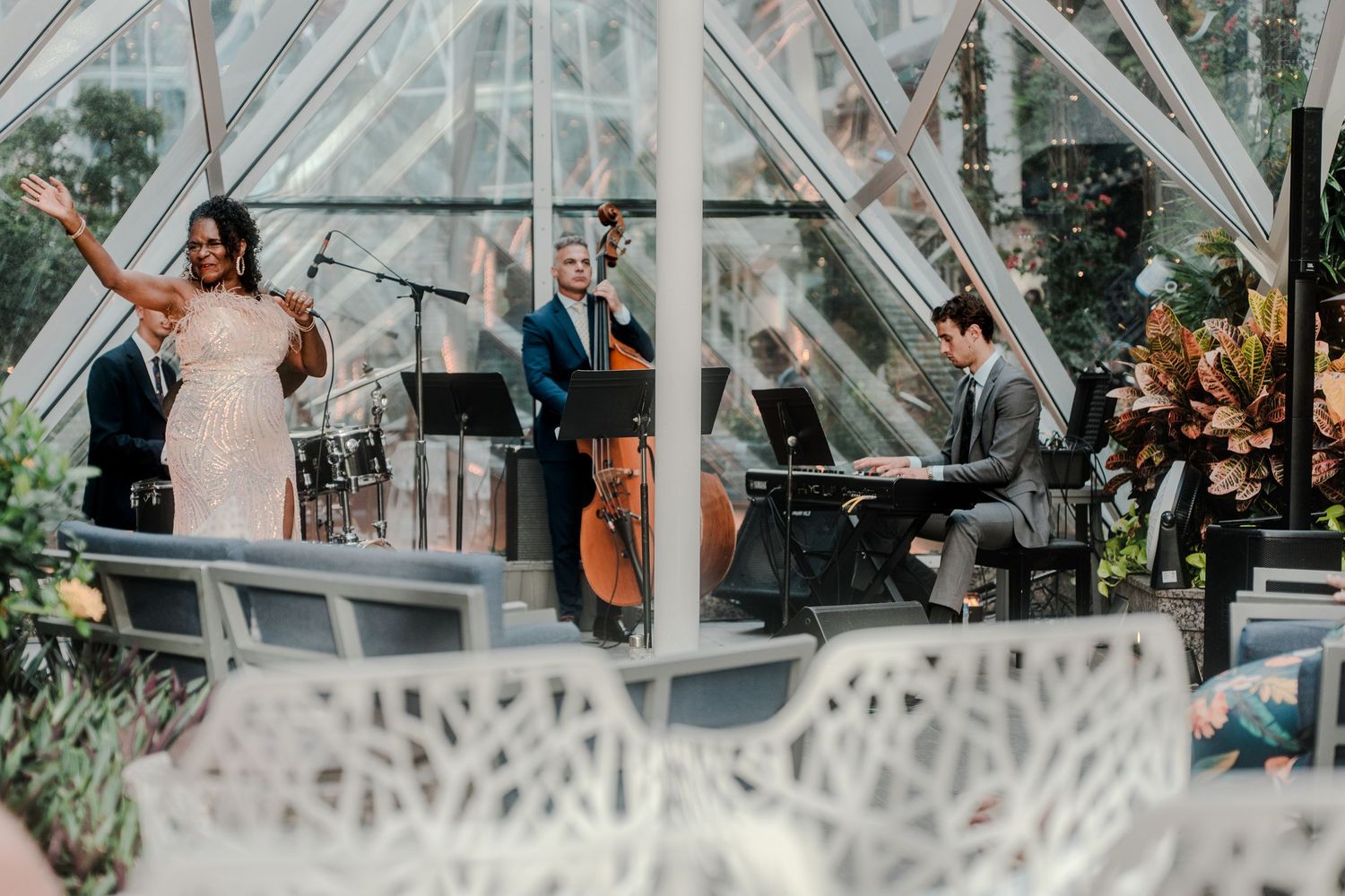 A live band with vocalist performs in a stunning glass greenhouse venue with white decorative furniture.
