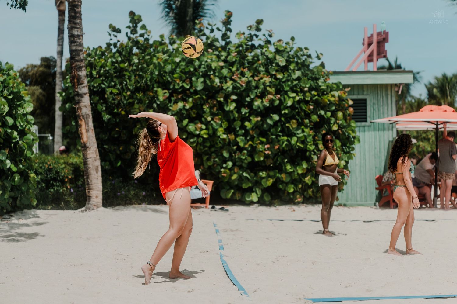 Woman in red shirt playing beach volleyball on white sand with palm trees and umbrellas in background.