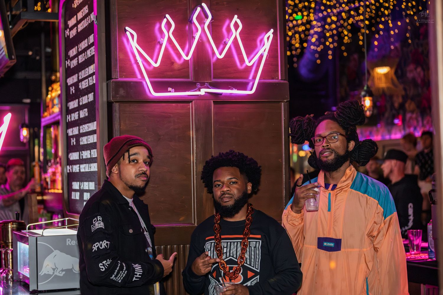 Three men posing together under a pink neon crown sign in a vibrant nightclub.