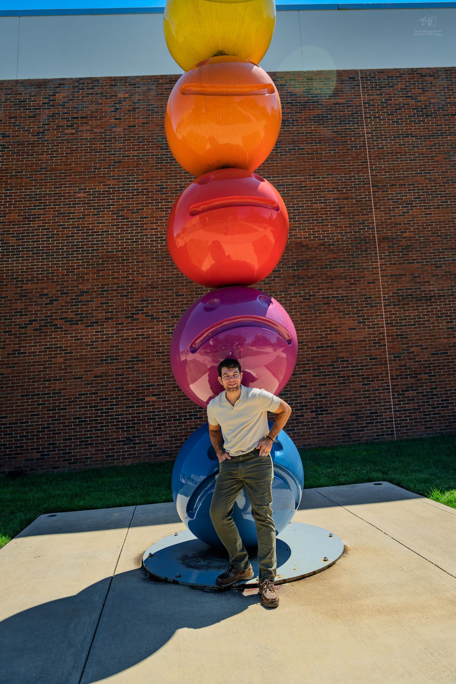 Man in white shirt leaning against tall multicolored stacked spheres sculpture outside brick wall.