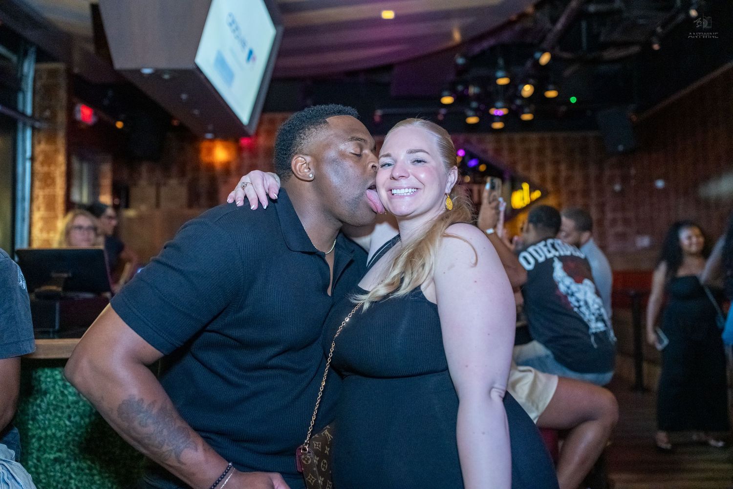 A man gives a woman a kiss on the cheek as they pose together at a crowded bar.