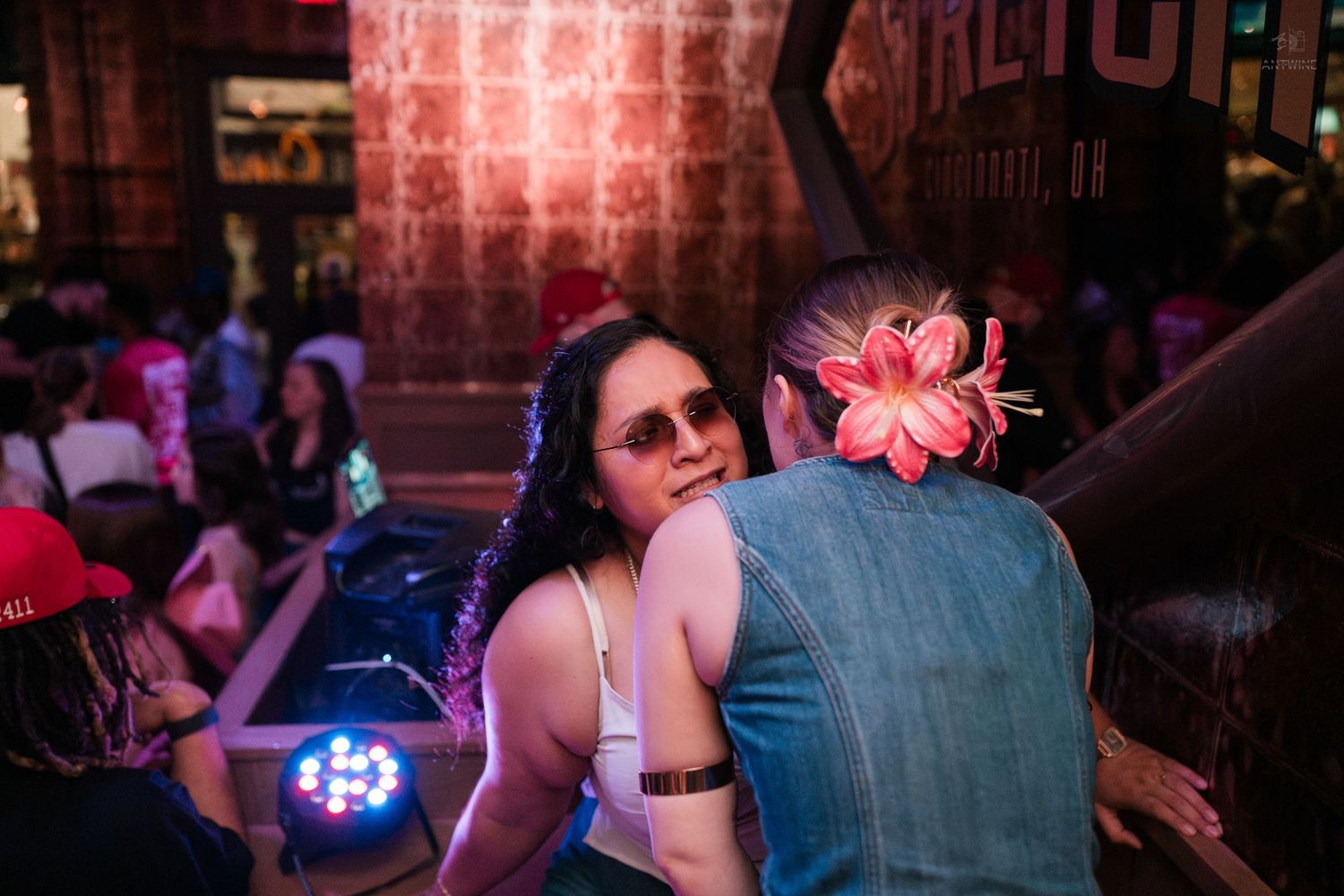 Two women talking closely at a nightclub event with colorful LED lighting nearby.