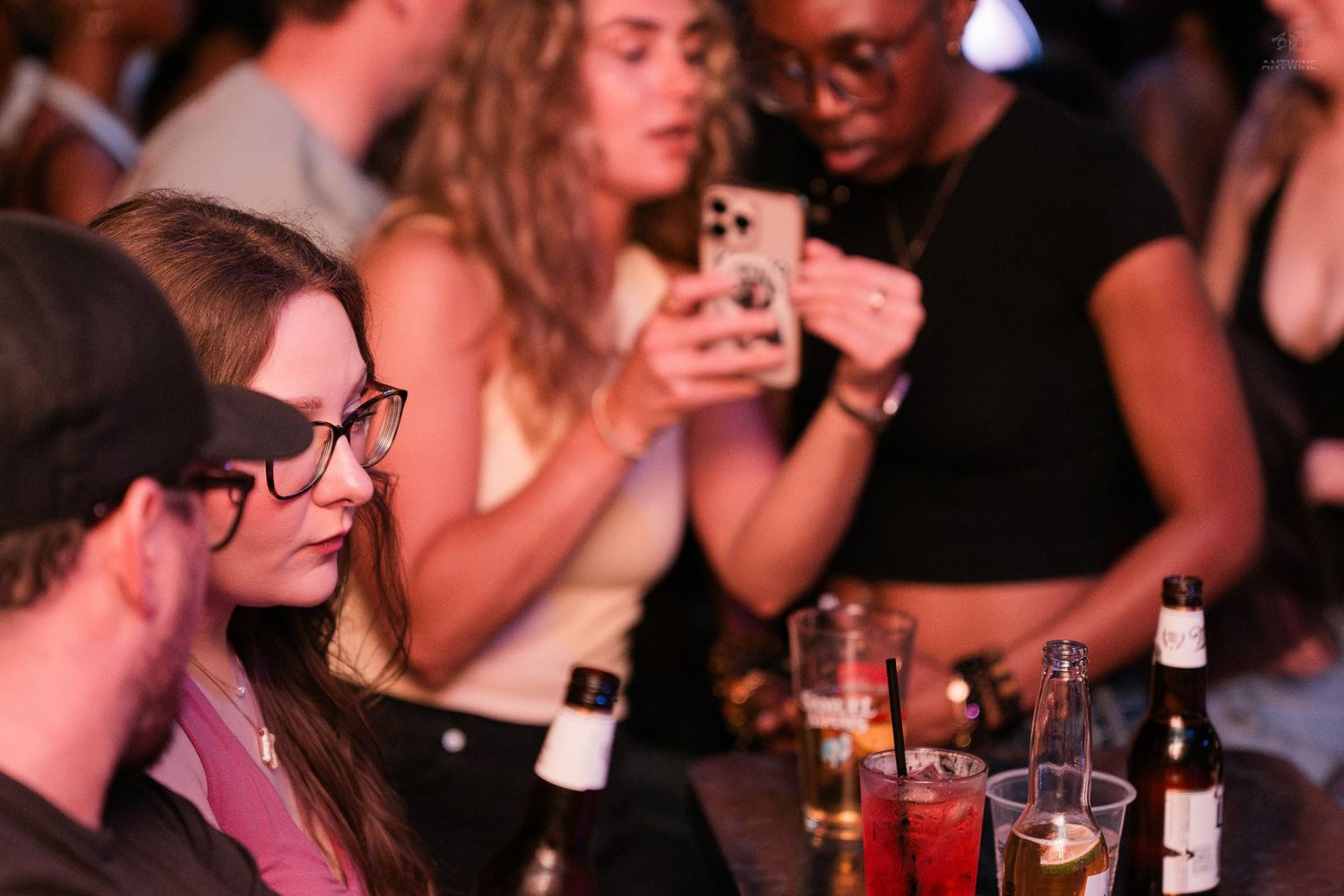 Young woman with glasses sitting at a bar table surrounded by drinks and people.