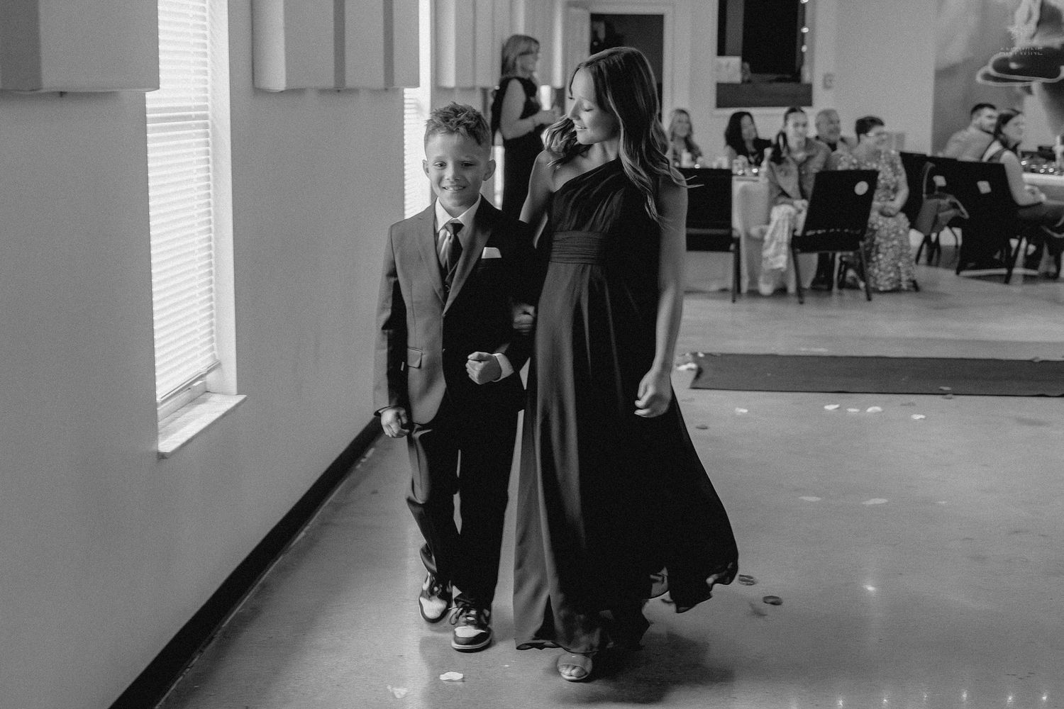 Young ring bearer walks with bridesmaid in black gown down wedding aisle in black and white photo.