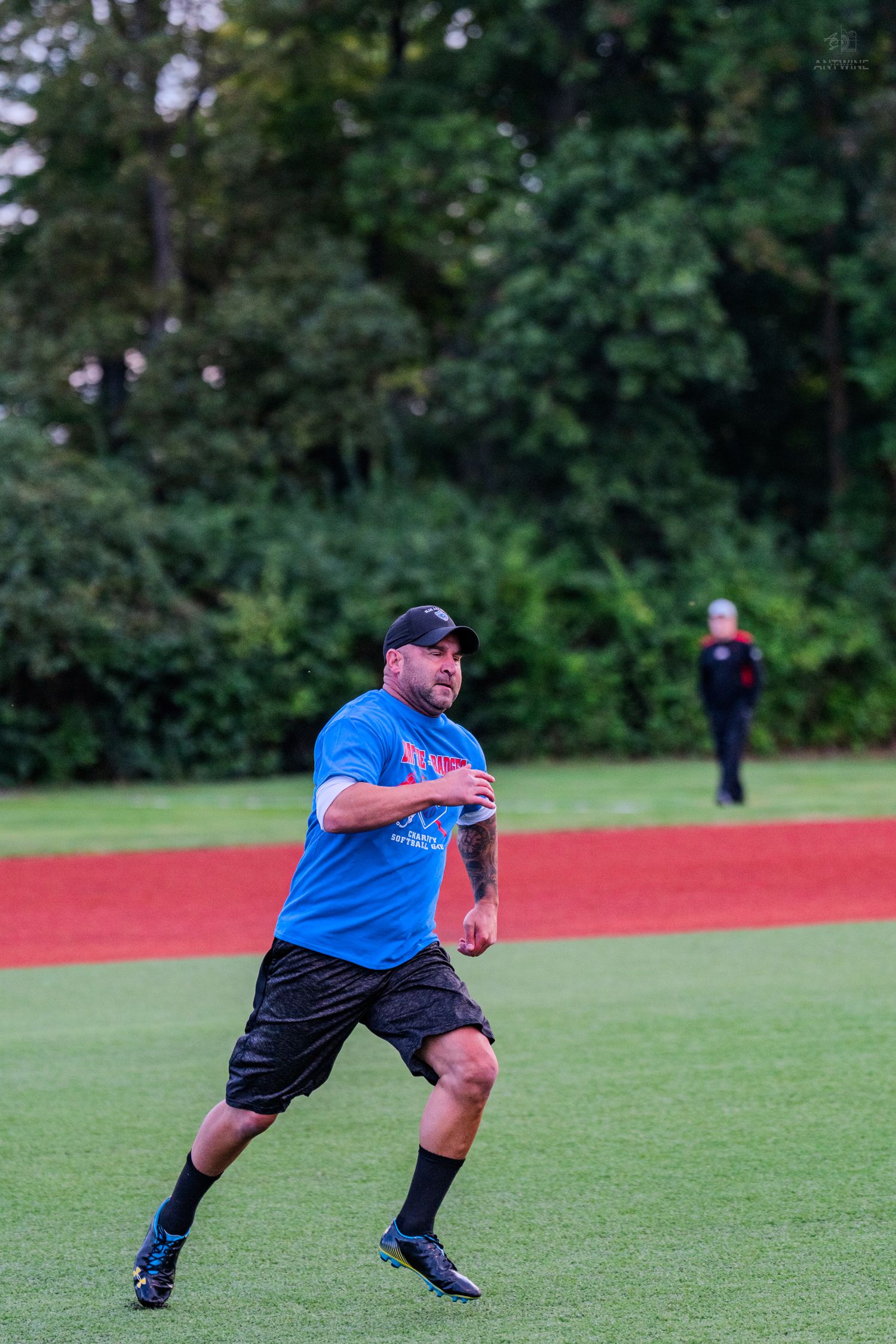 Man in blue shirt and black shorts running on a softball field during an outdoor recreational game at dusk.