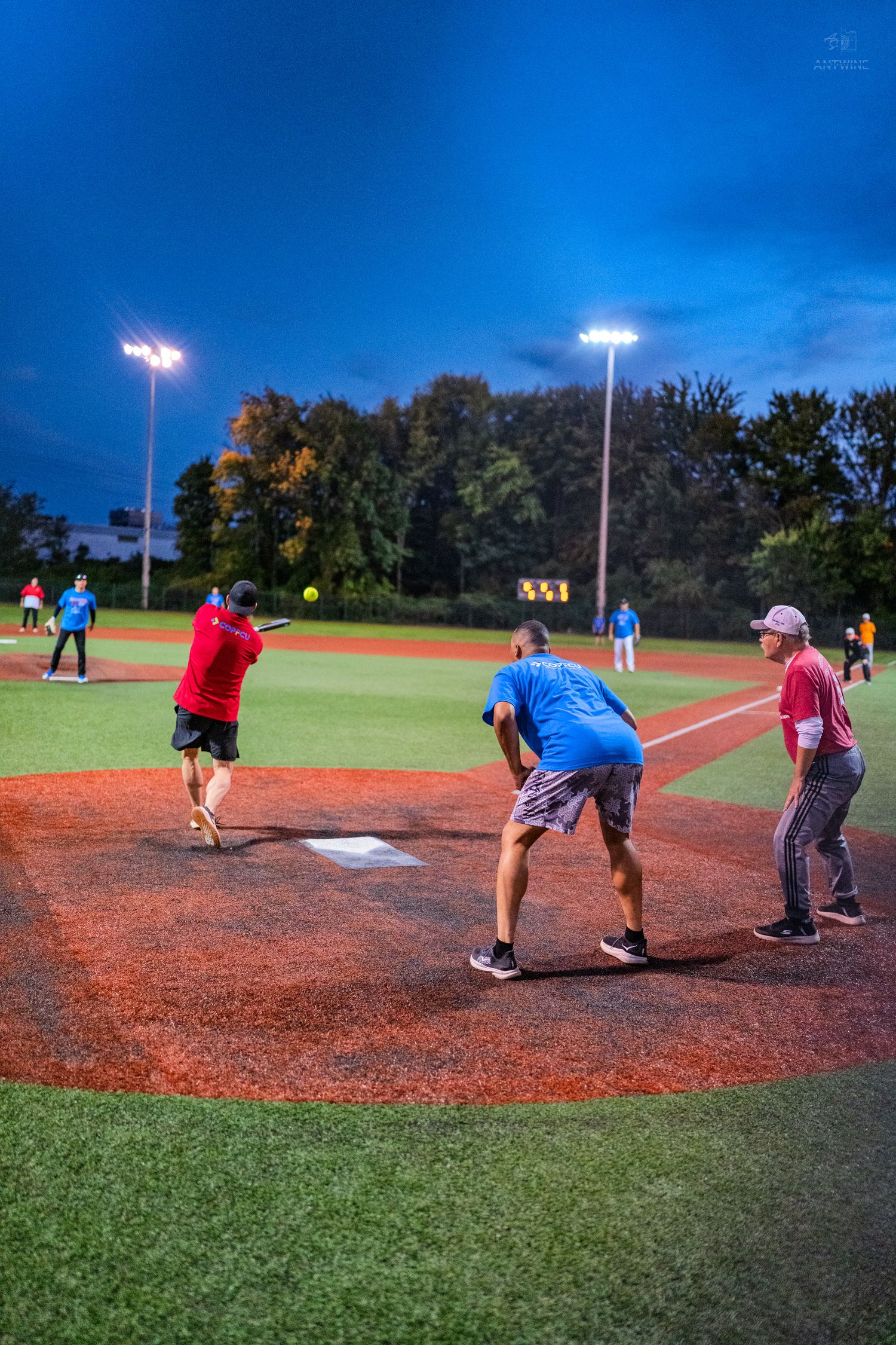 Players compete in a nighttime softball game under bright field lights on a red turf diamond.