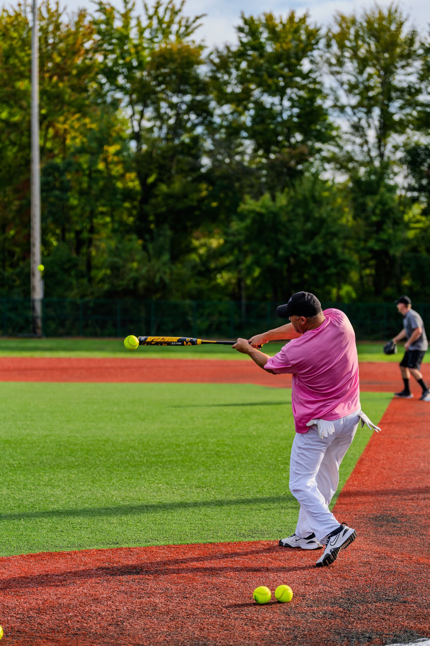 A batter in a pink shirt swings at a softball on a red clay field during an outdoor practice session.