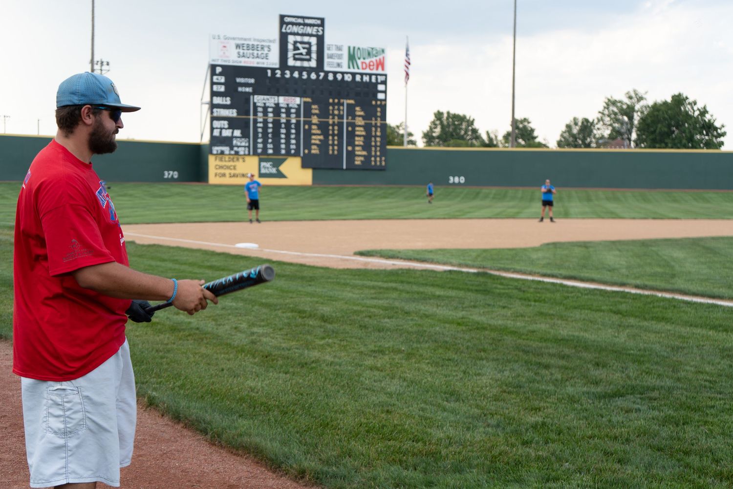 Baseball player holding a bat watches teammates practice on a field with a large scoreboard behind him.