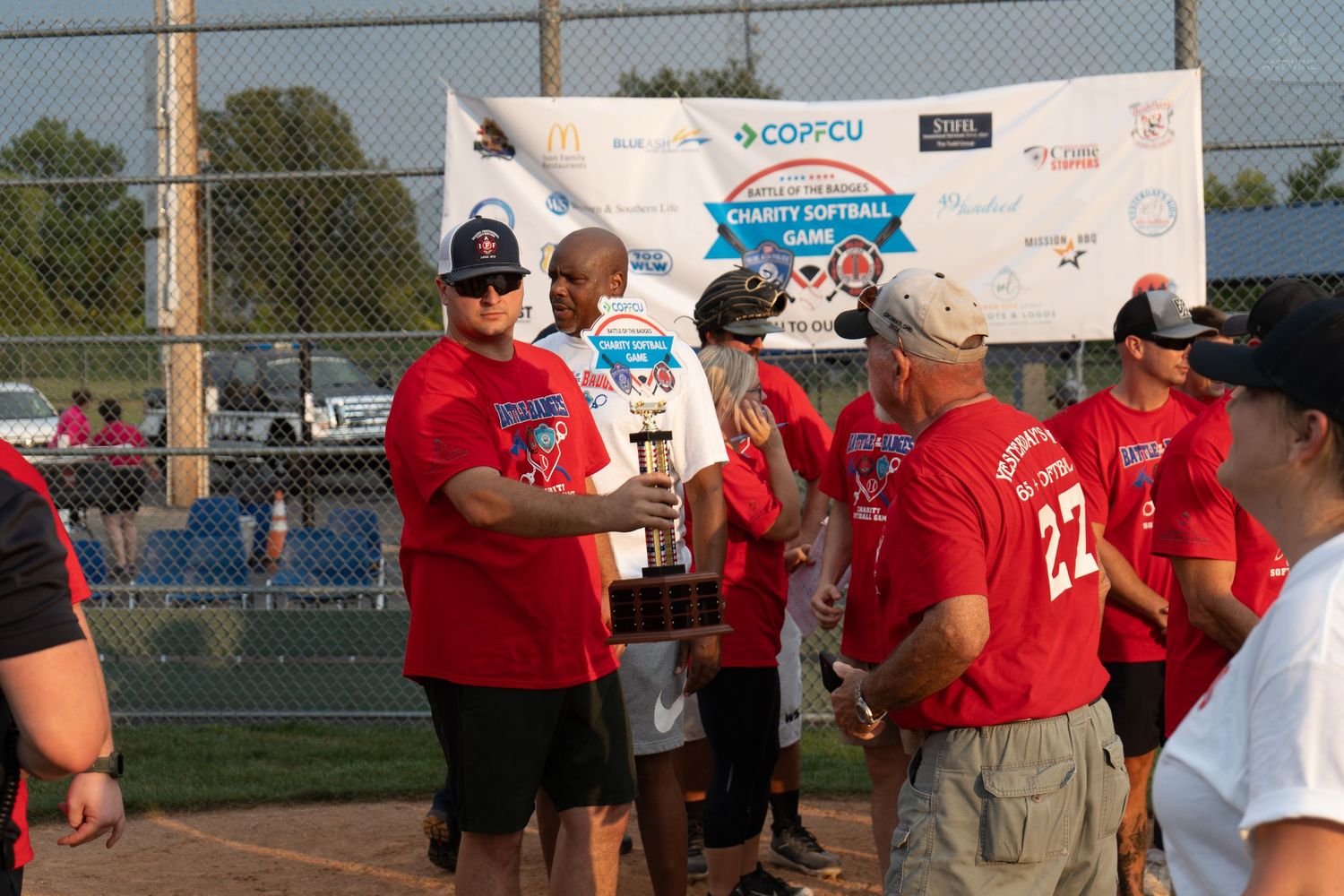 Players in red uniforms present a trophy at a COPCU charity softball game event.