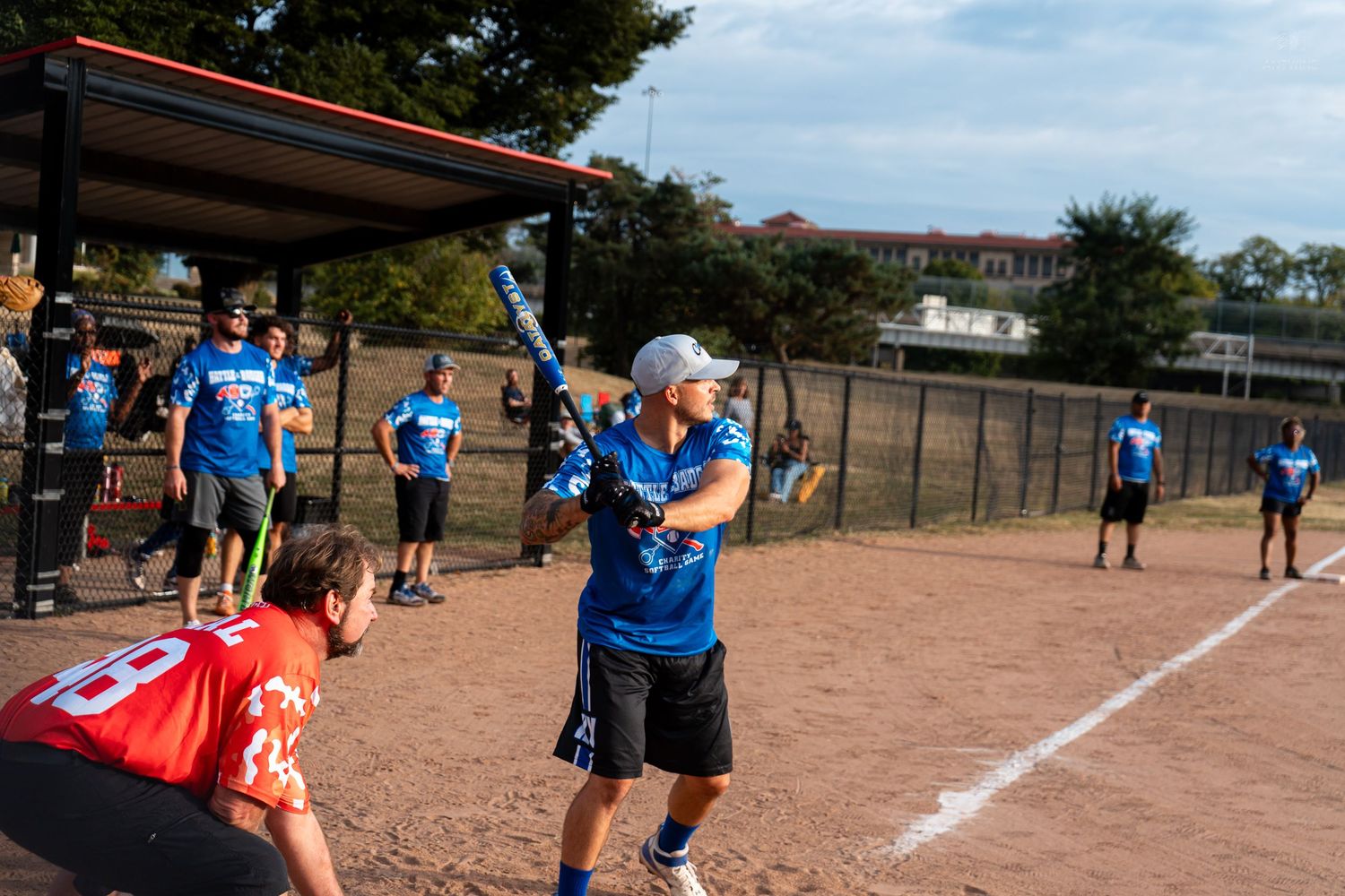 A batter swings during an outdoor softball game with teammates in blue shirts watching from the dugout.