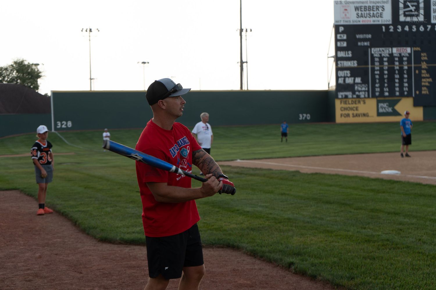 A man in a red shirt holding a blue bat at a baseball field with a scoreboard in the background.