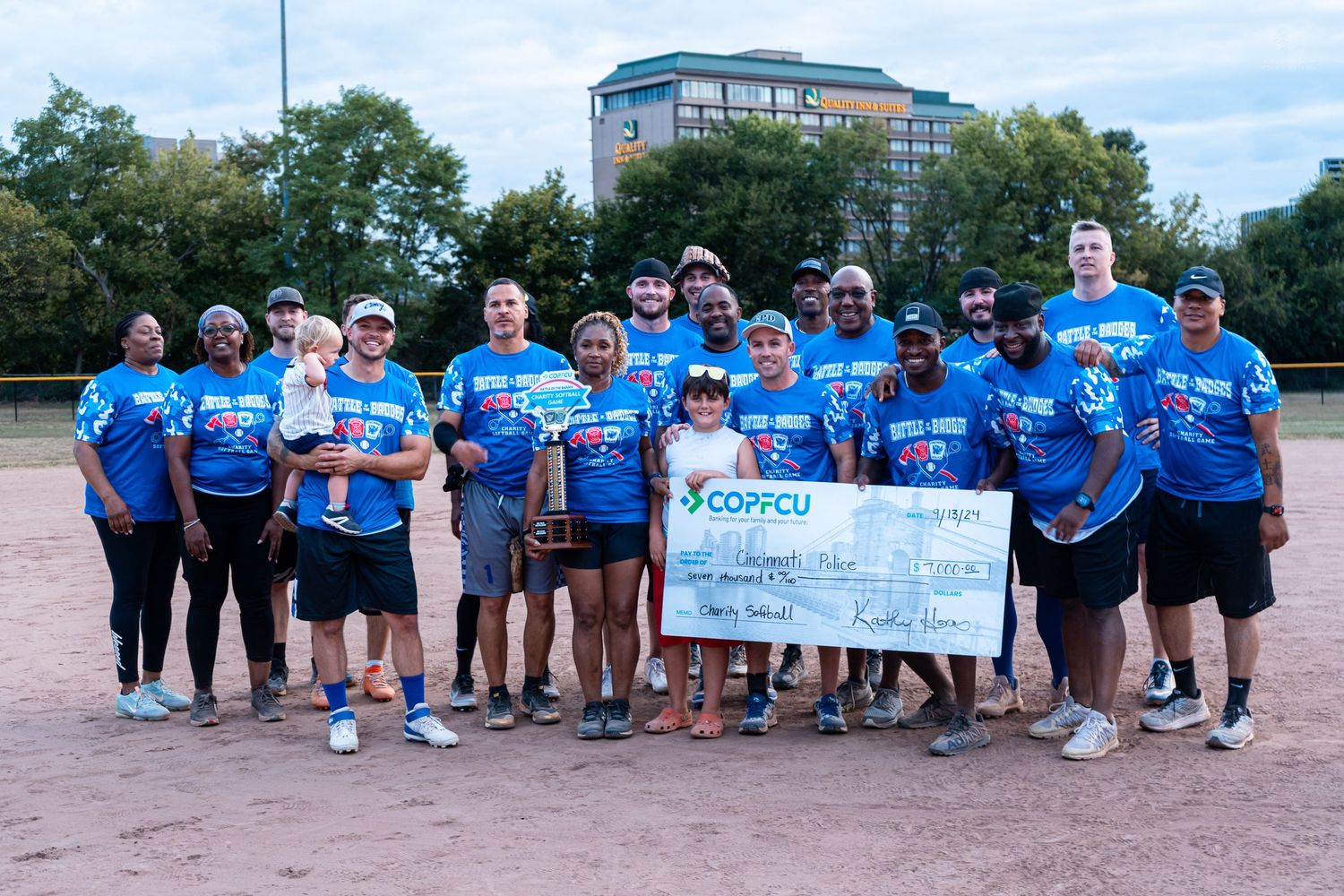 Winning softball team poses with trophy and COPFCU sponsor check on outdoor dirt field.