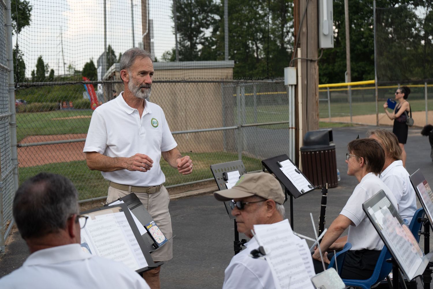 Outdoor band rehearsal at a baseball field with a conductor leading musicians reading sheet music on stands.