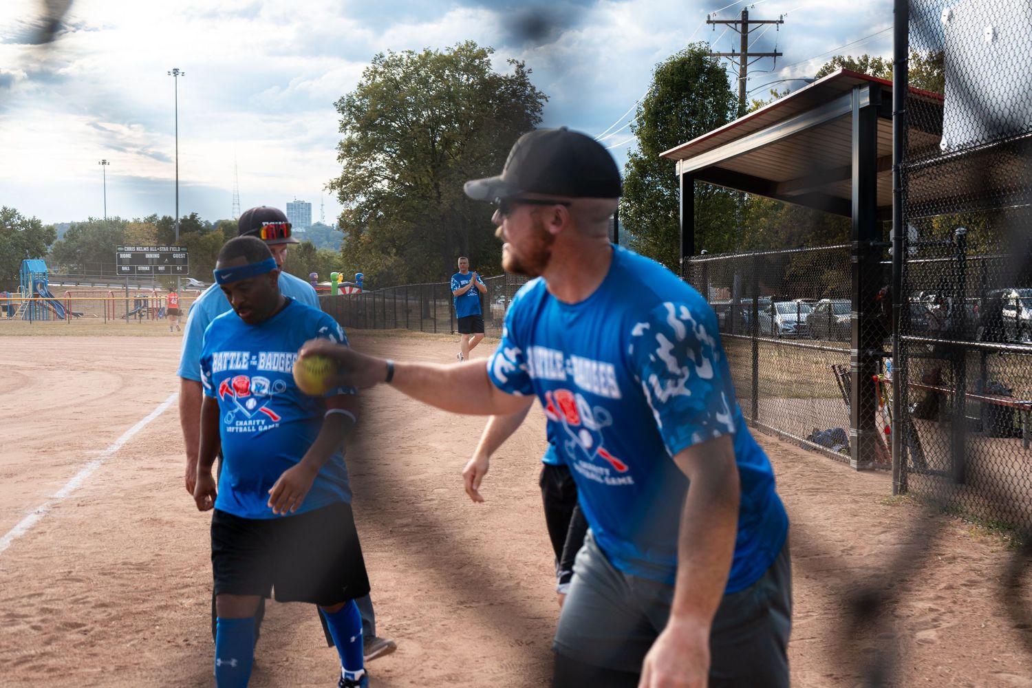 Players in blue jerseys compete in a recreational softball game on an outdoor diamond on a sunny day.