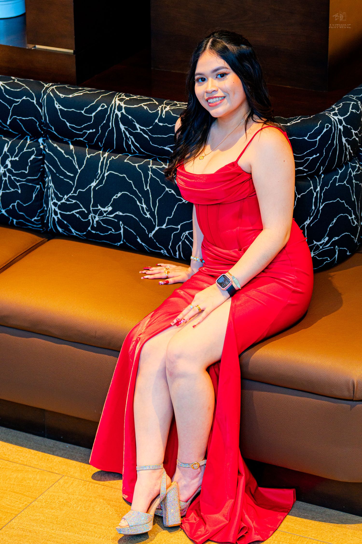 Young woman in red slit gown and heels sitting on brown leather sofa smiling at camera.