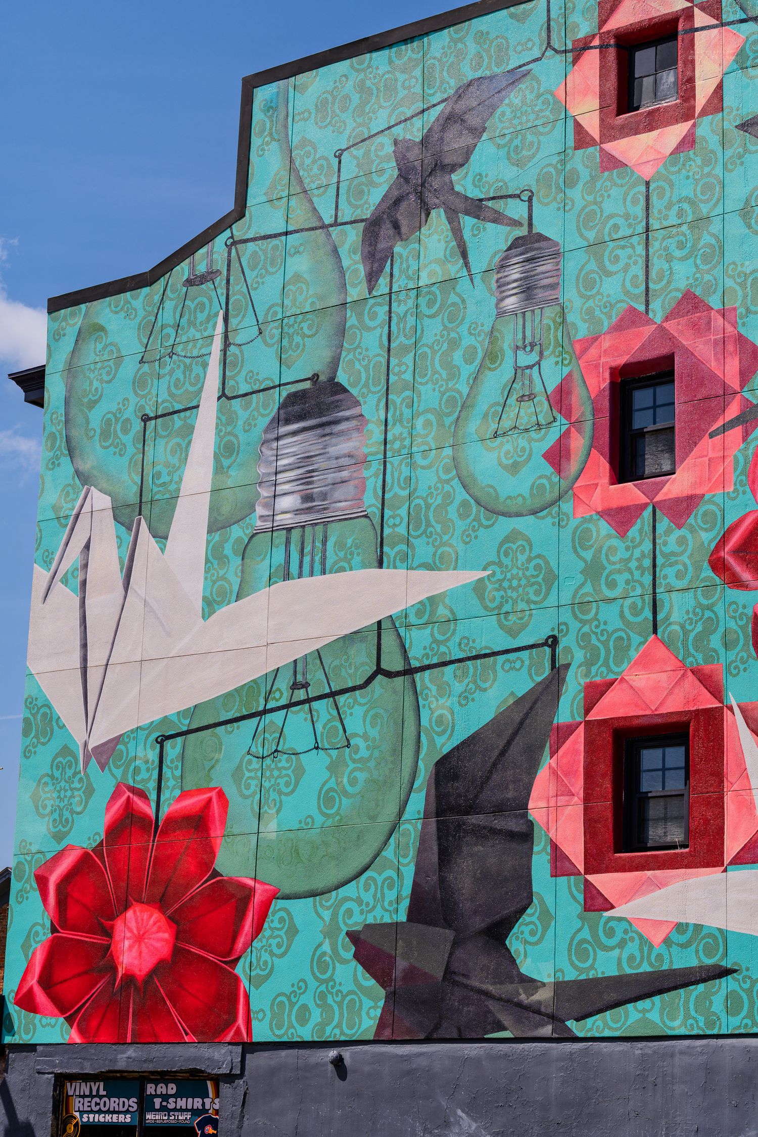 Vibrant teal building mural featuring red geometric flowers, light bulbs, origami birds, and decorative patterns.