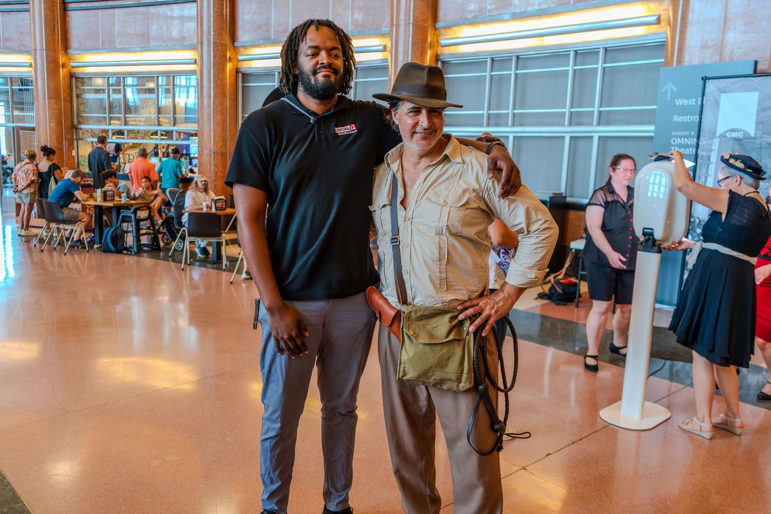 Two men pose together indoors, one in a black polo shirt and one dressed as Indiana Jones with a whip.