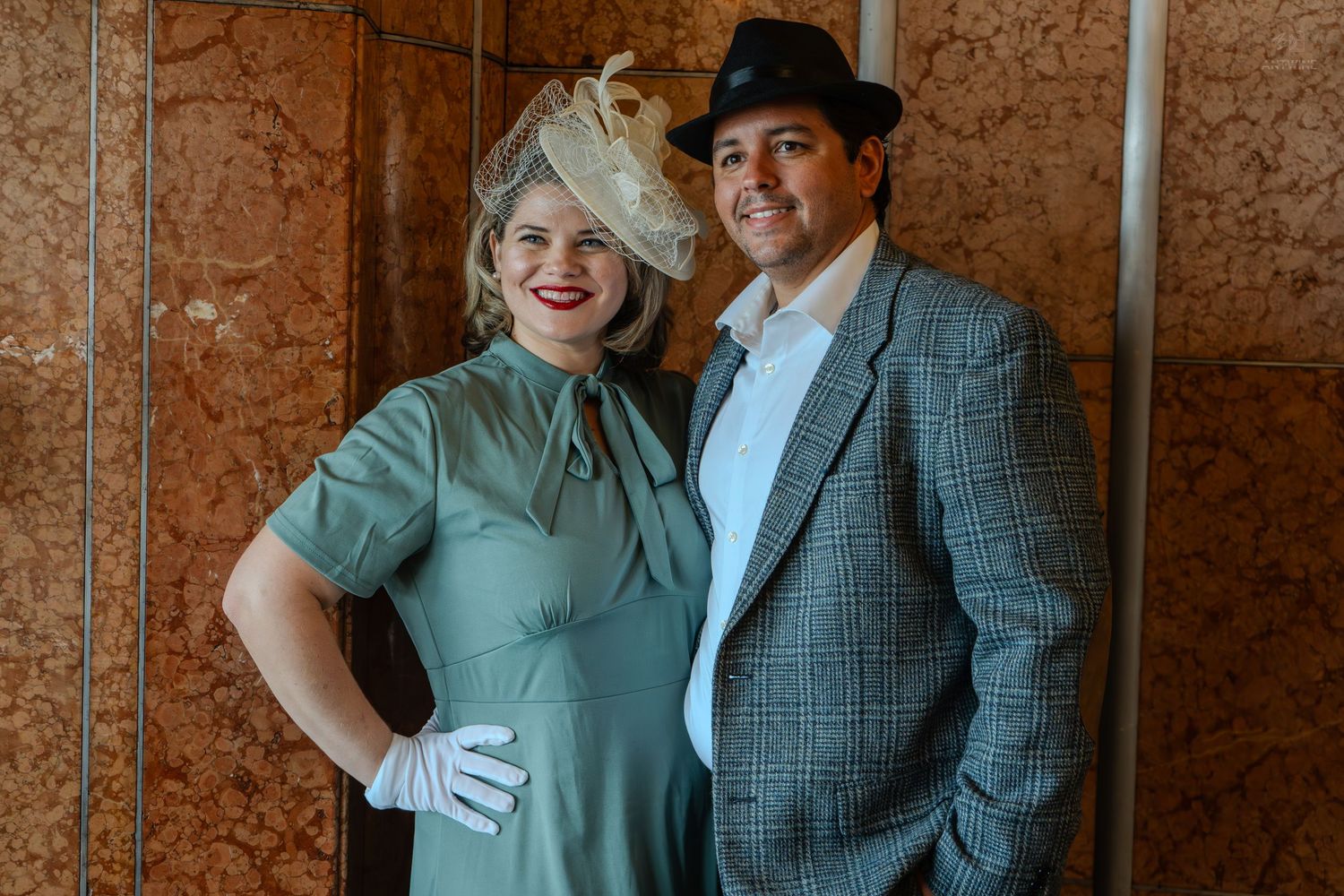 Couple in vintage 1940s attire, woman in green dress with white gloves, man in plaid suit and fedora hat.