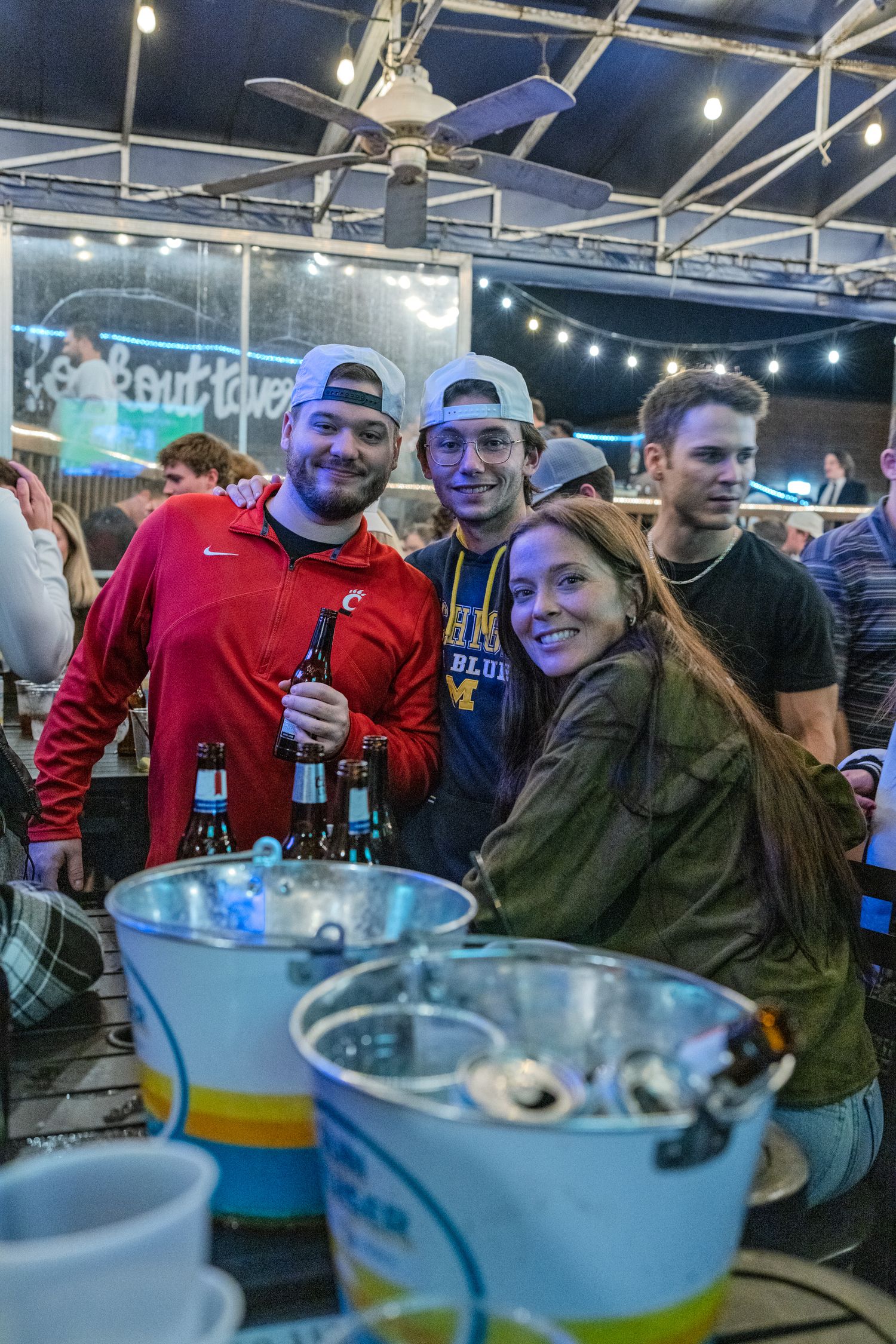 Three friends smile and pose with beer bottles at a lively outdoor bar patio at night.