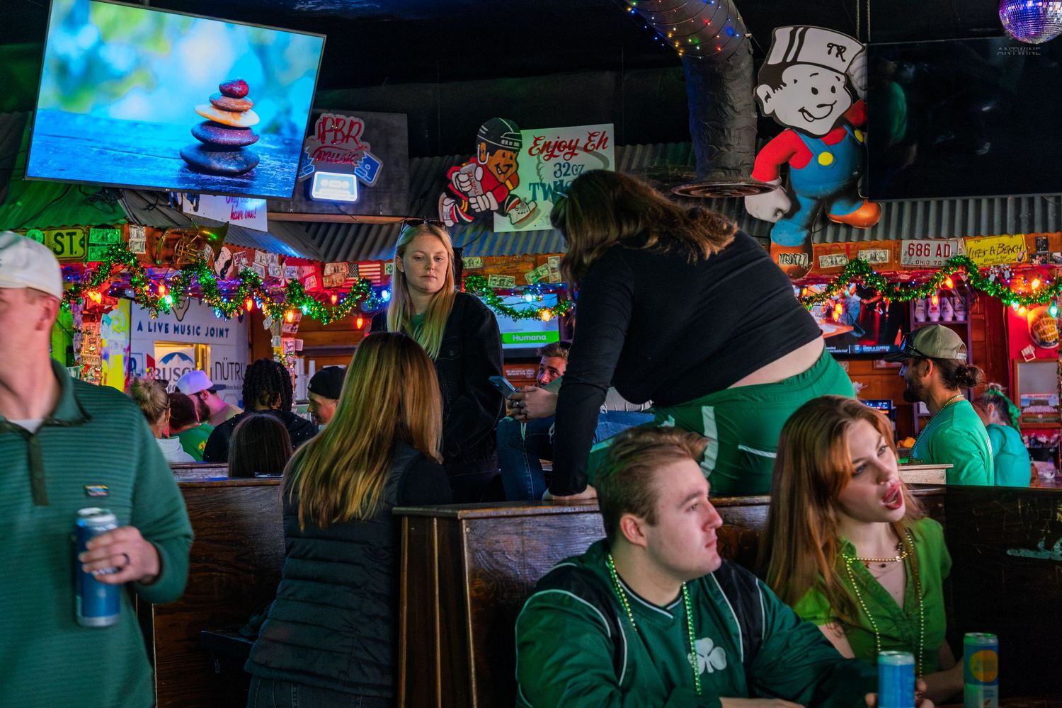 Busy Irish bar with patrons in green attire, a waitress serving tables, TVs, and festive St. Patrick's Day decorations.