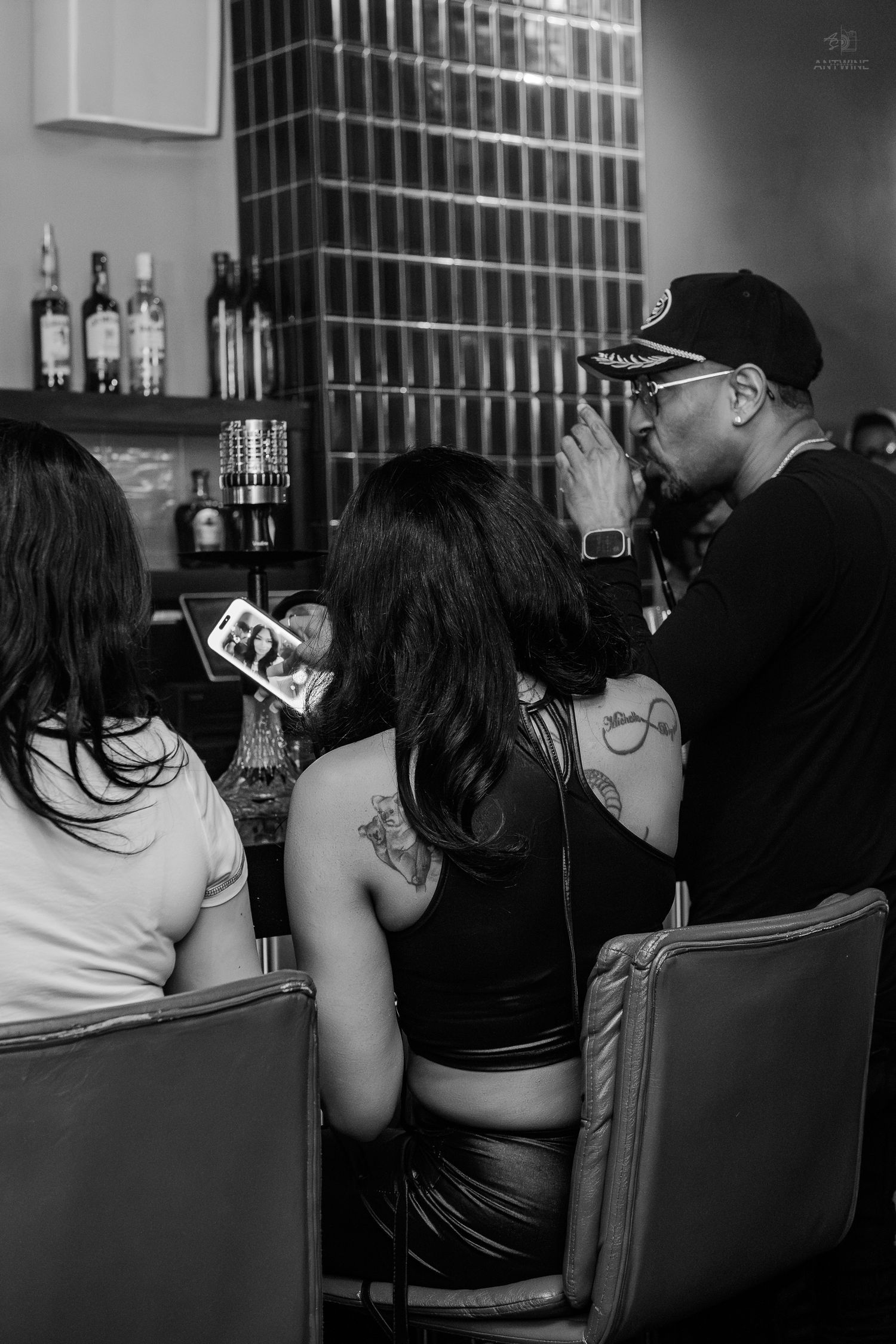 Black and white photo of tattooed women and a man at a bar, one holding a phone, liquor bottles in background.