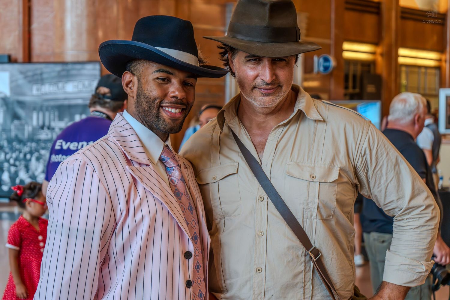 Two men in vintage costumes pose smiling, one in pinstripe suit with hat, other in safari outfit with fedora.