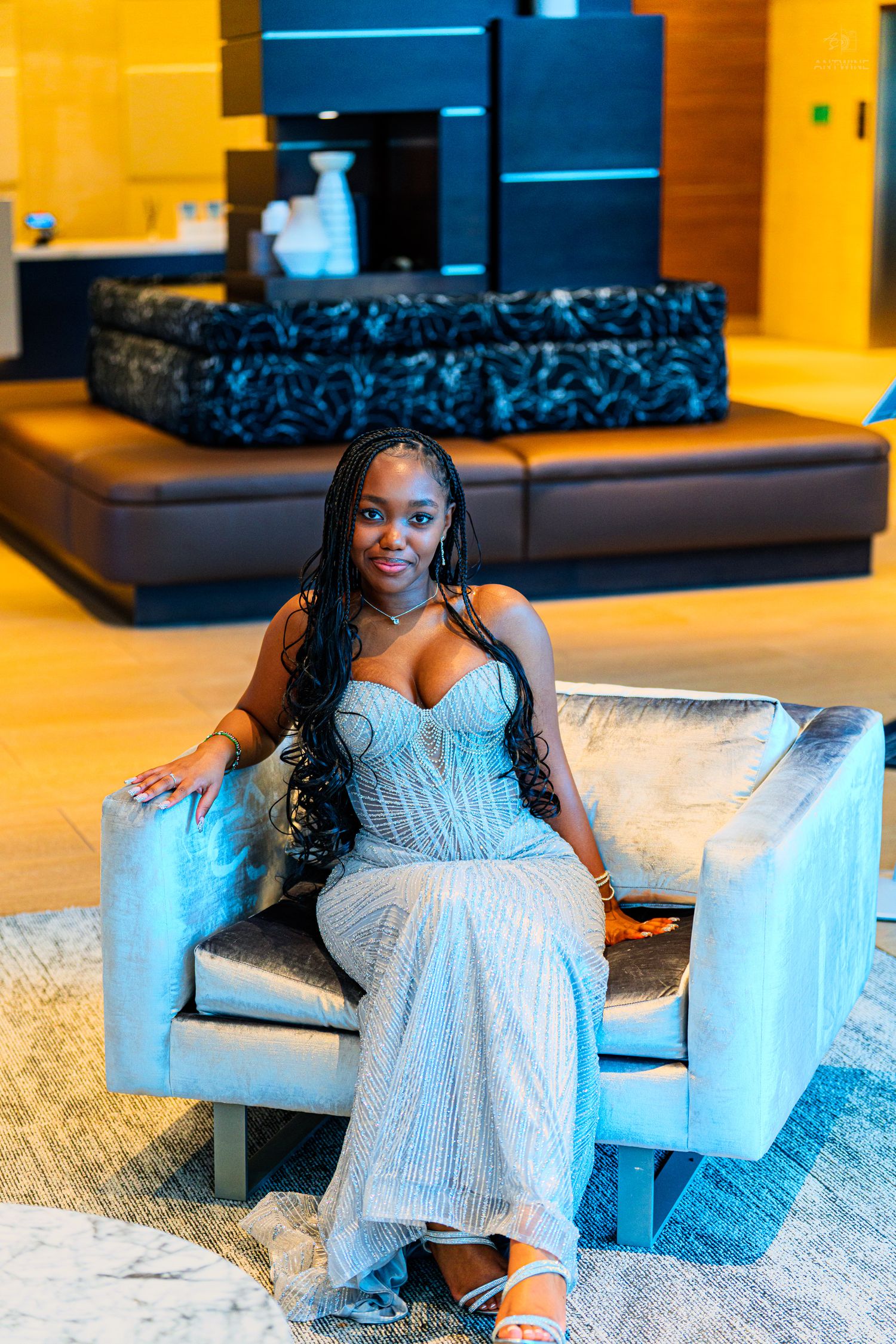 Woman in silver sequin gown seated in modern hotel lobby chair smiling confidently.