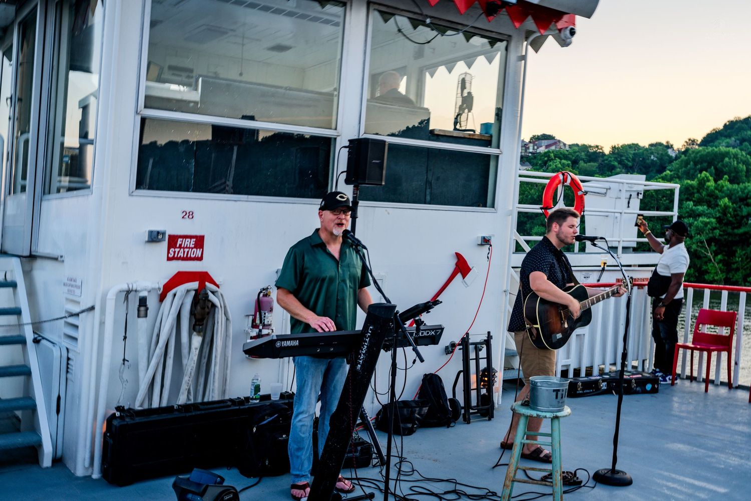 Live band performing outdoors on a riverboat deck at dusk with two musicians playing keyboard and guitar.