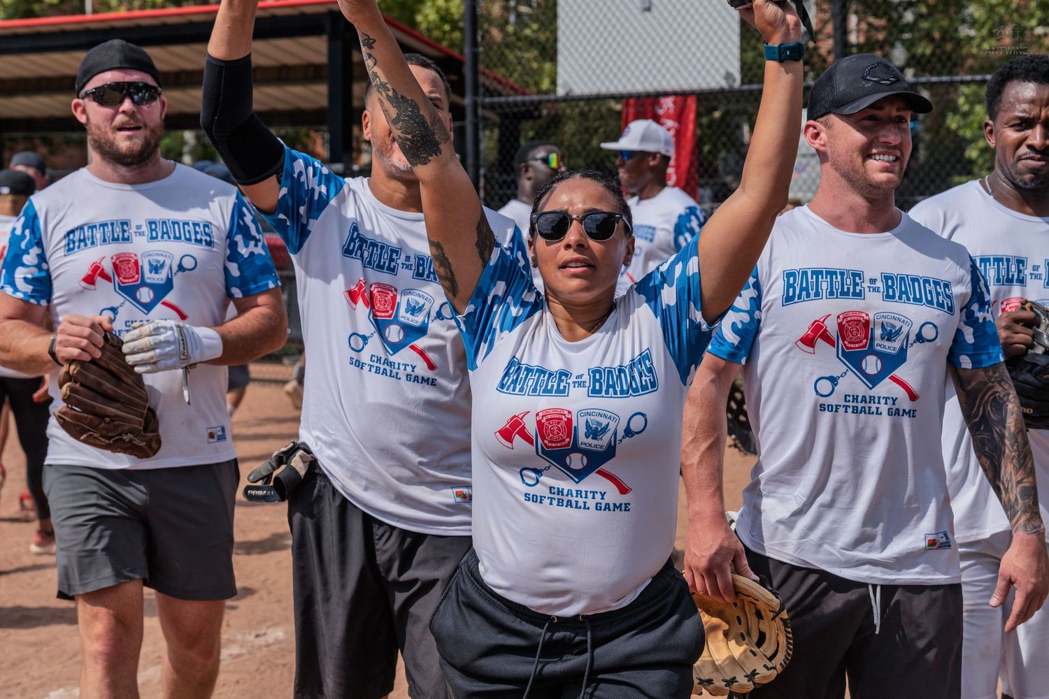 Softball players in Battle of the Badges charity game uniforms celebrate with raised arms on the field.