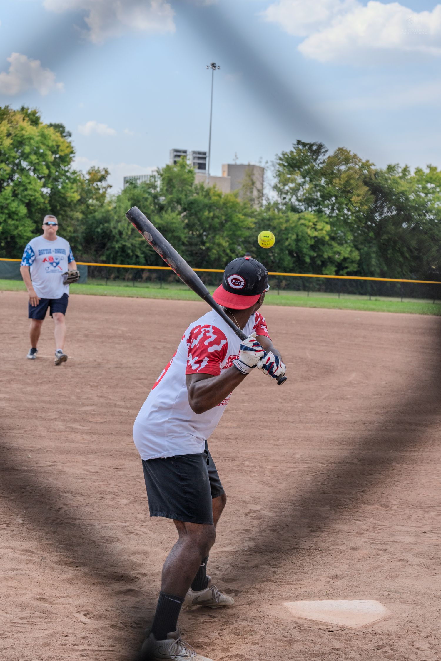 Softball batter swinging at a yellow ball during a recreational game on a dirt field with players in the background.