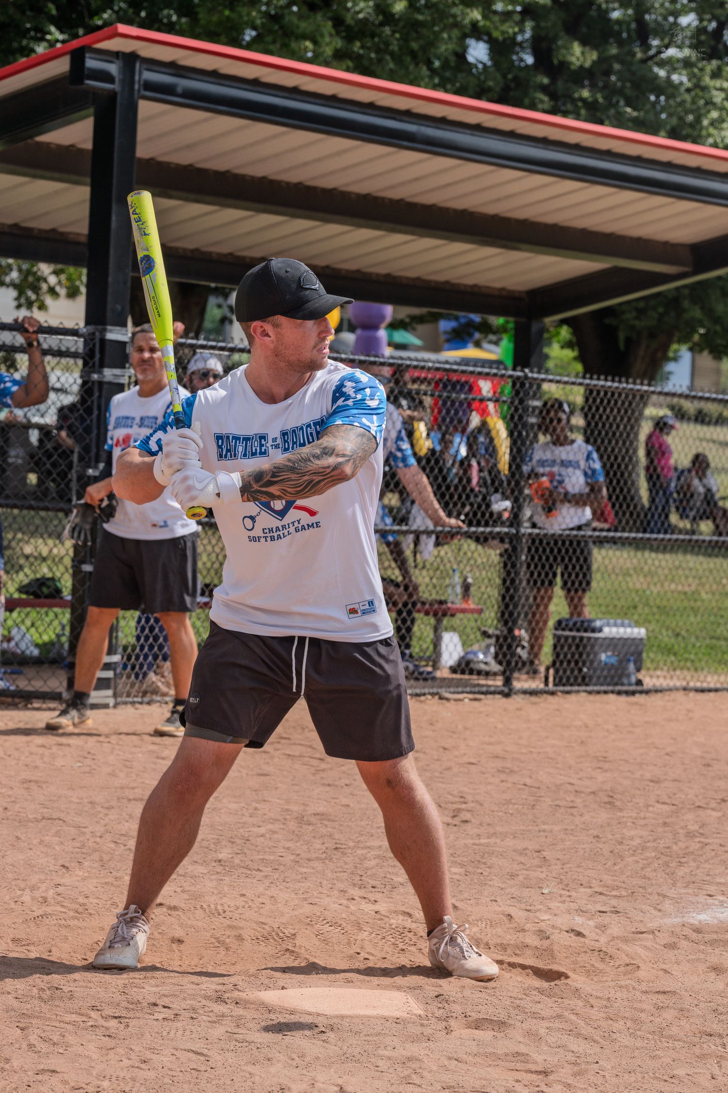 A tattooed man in a white team jersey holds a yellow bat ready to swing at a softball game.