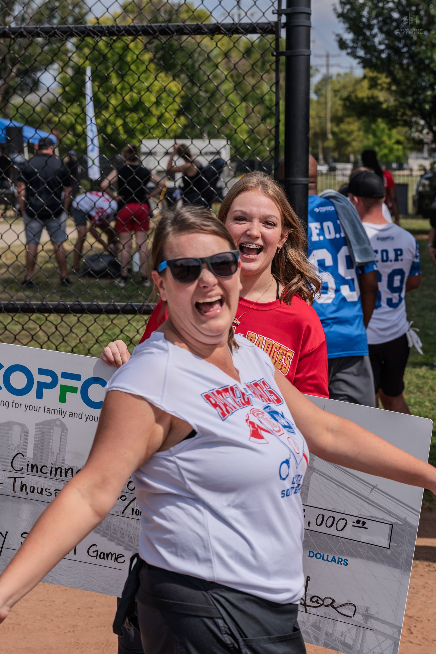Two women celebrate holding a COPFCU charity softball game check for Cincinnati Police at an outdoor event.